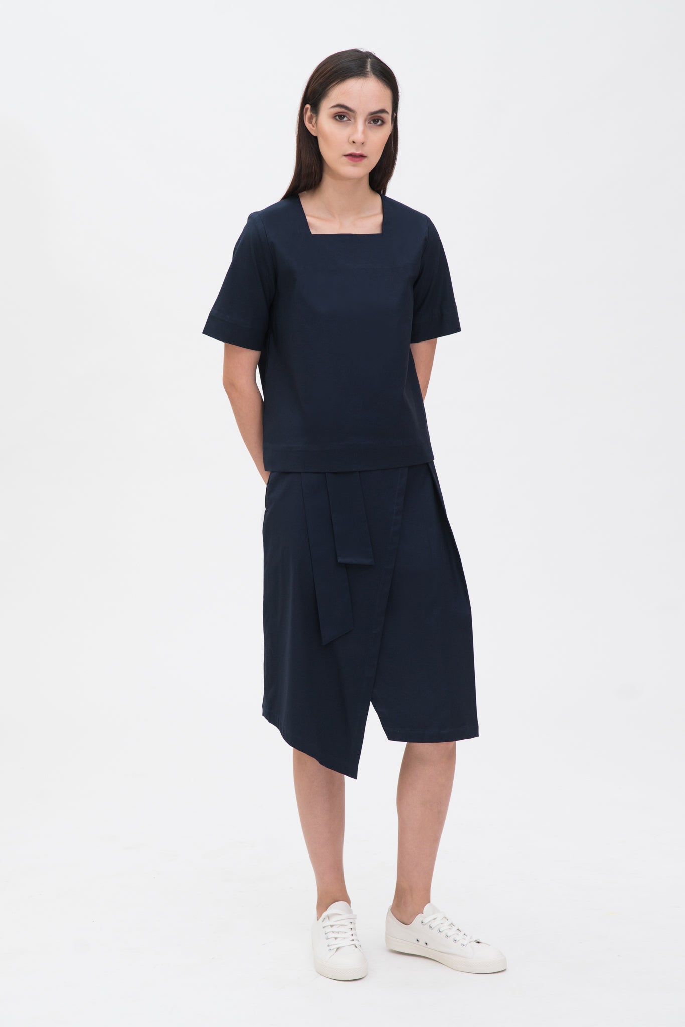 A woman with long dark hair is wearing a navy t-shirt and asymmetric navy skirt  with white shoes