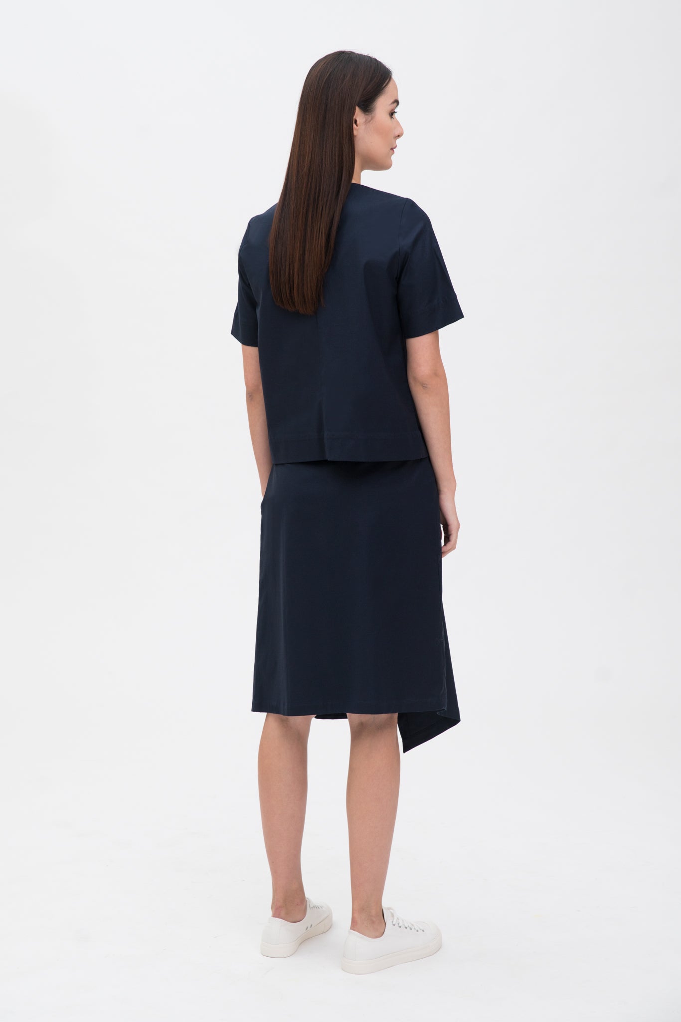 A woman with long dark hair is wearing a navy t-shirt and asymmetric navy skirt  with white shoes