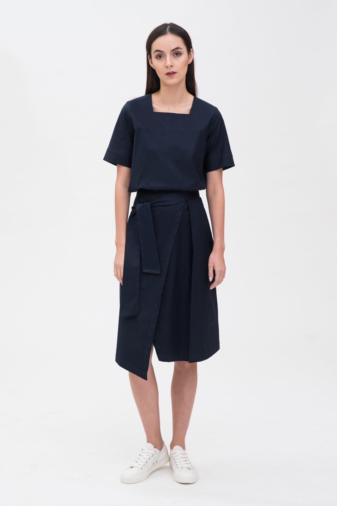 A woman with long dark hair is wearing a navy t-shirt and asymmetric navy skirt  with white shoes