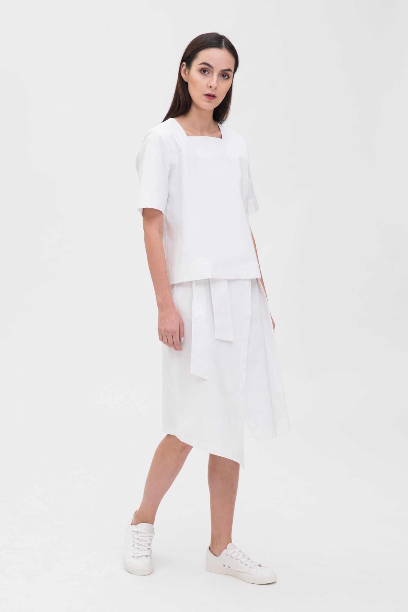 A woman with long dark hair is wearing a navy t-shirt and asymmetric white skirt  with white shoes