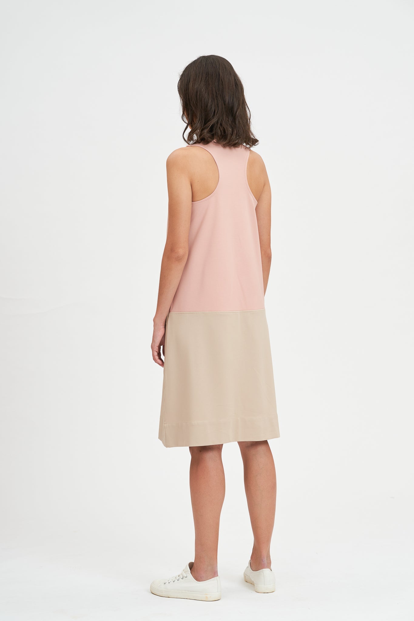 A woman with shoulder length brown hair is wearing a pale pink and nude knee length dress. She also has white tennis shoes on.