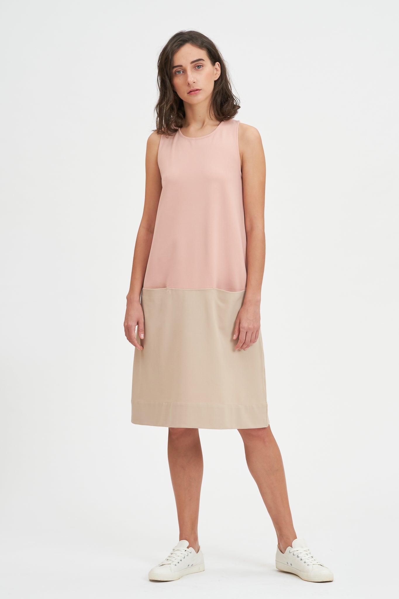 A woman with shoulder length brown hair is wearing a pale pink and nude knee length dress. She also has white tennis shoes on.