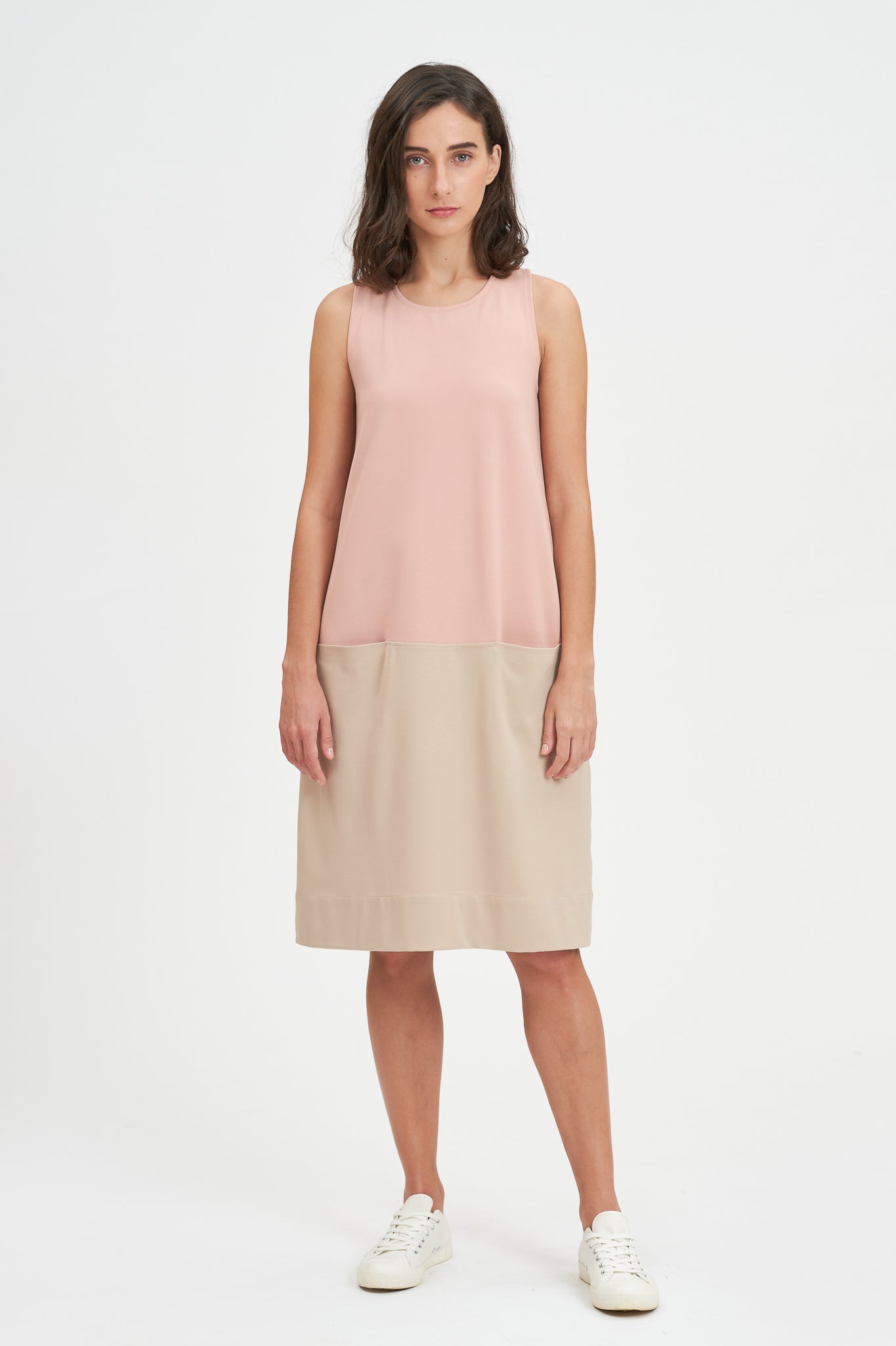 A woman with shoulder length brown hair is wearing a pale pink and nude knee length dress. She also has white tennis shoes on.