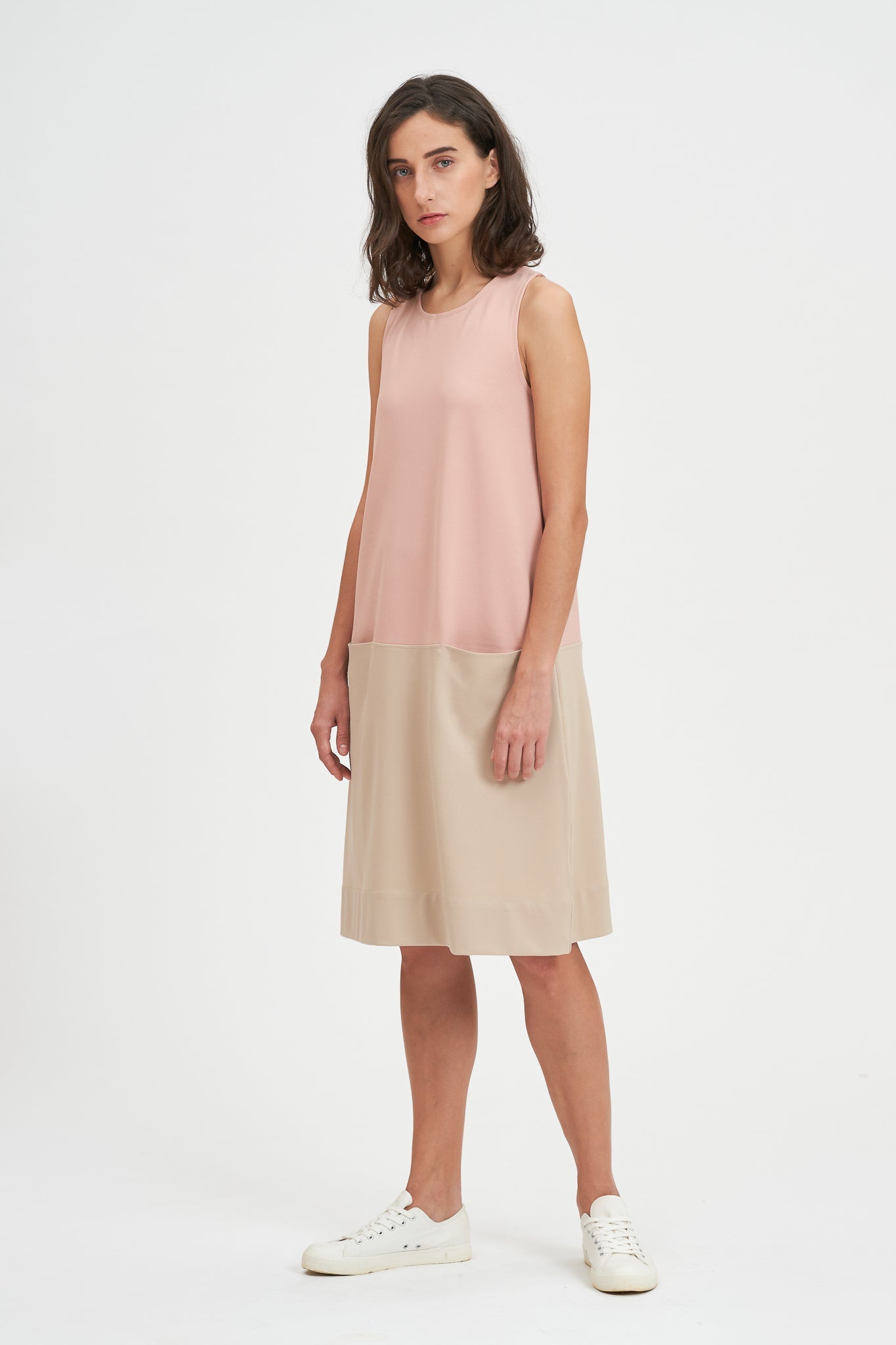 A woman with shoulder length brown hair is wearing a pale pink and nude knee length dress. She also has white tennis shoes on.