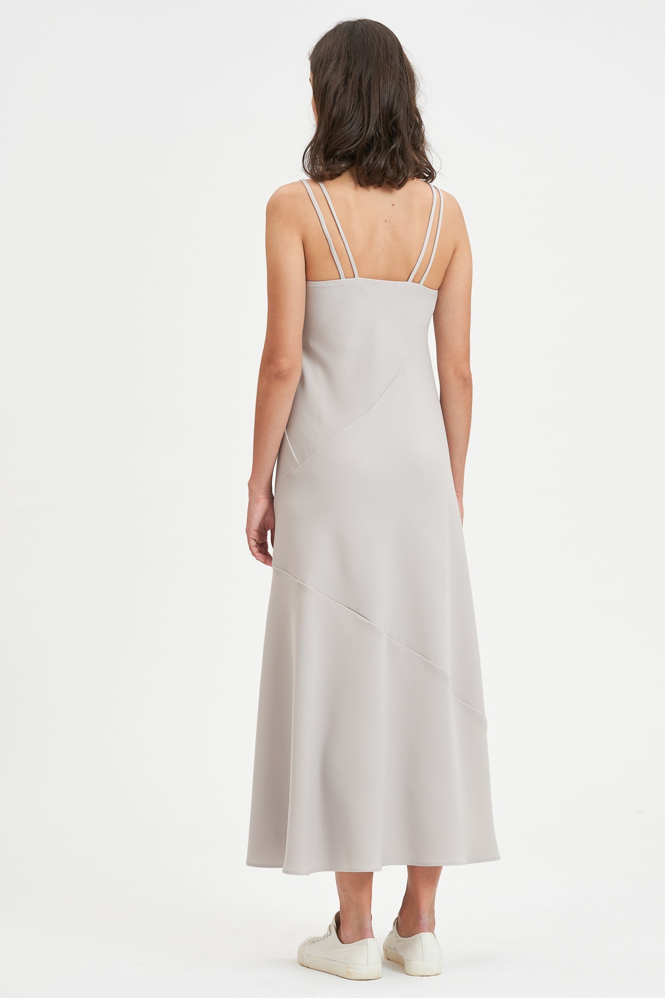 A woman with shoulder length hair is wearing an ankle length gray strappy dress. She is wearing white tennis shoes.