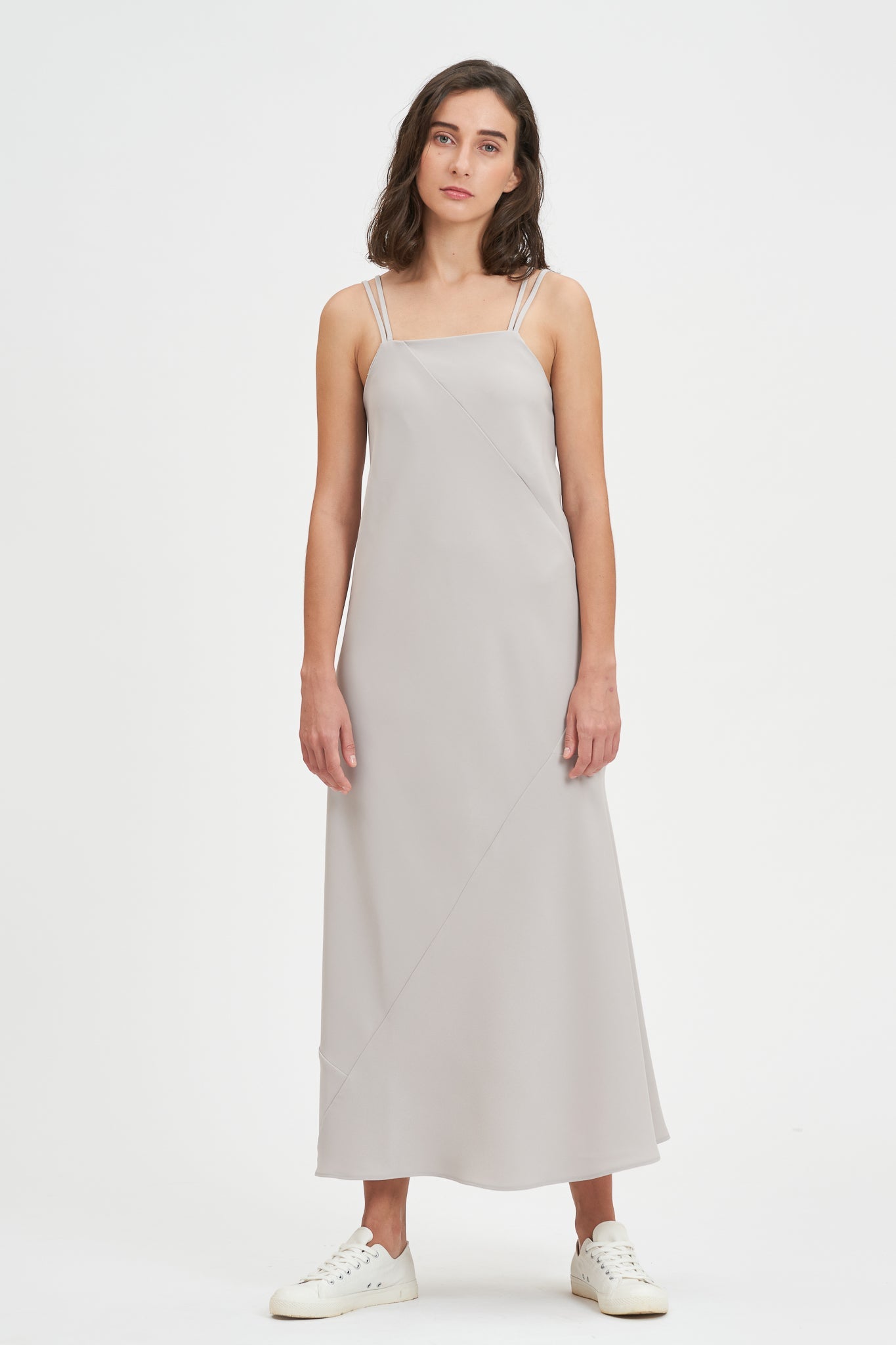 A woman with shoulder length hair is wearing an ankle length gray strappy dress. She is wearing white tennis shoes.