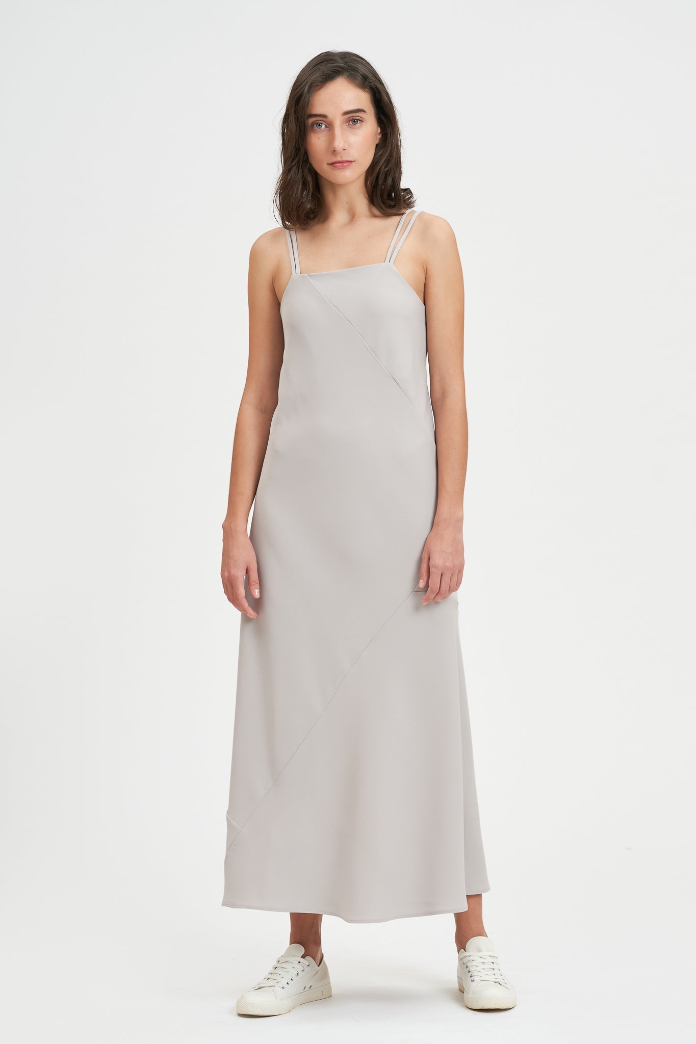 A woman with shoulder length hair is wearing an ankle length gray strappy dress. She is wearing white tennis shoes.