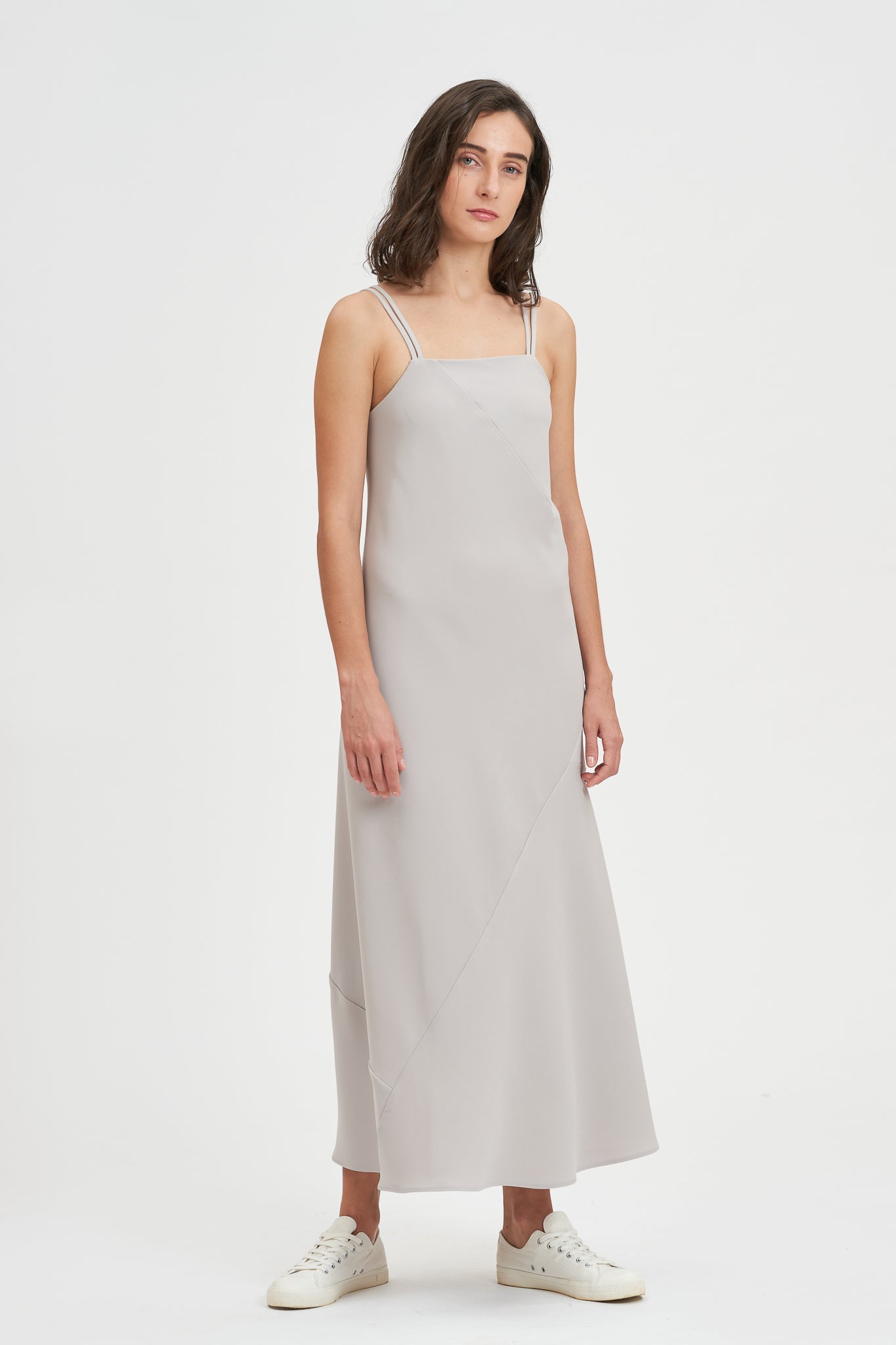 A woman with shoulder length hair is wearing an ankle length gray strappy dress. She is wearing white tennis shoes.