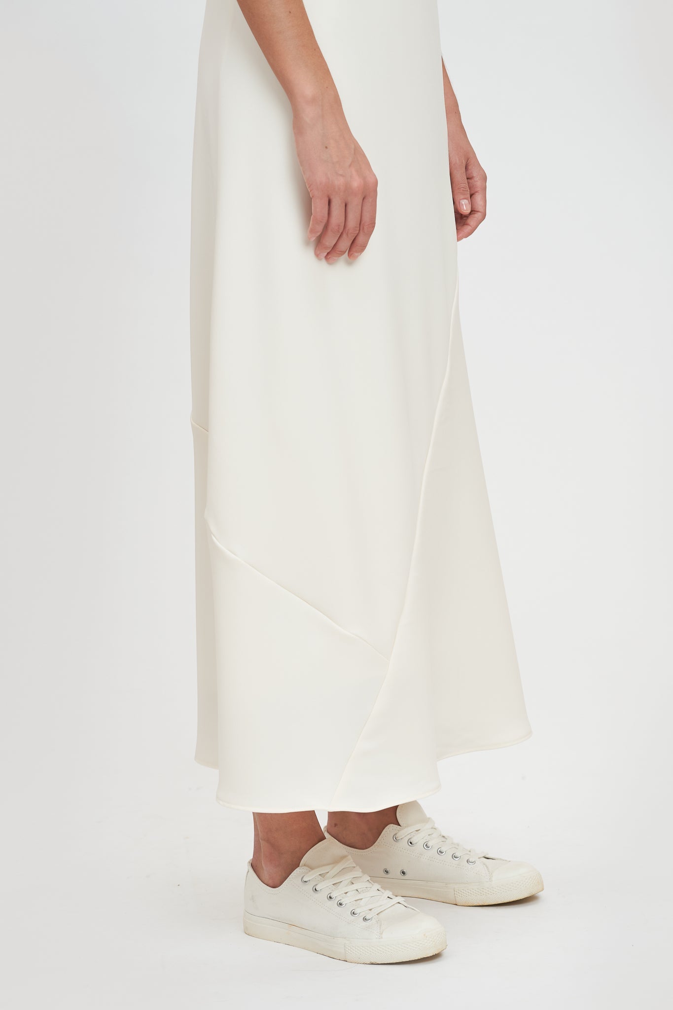 A woman with shoulder length hair is wearing an ankle length white scrappy dress. She is wearing white tennis shoes.