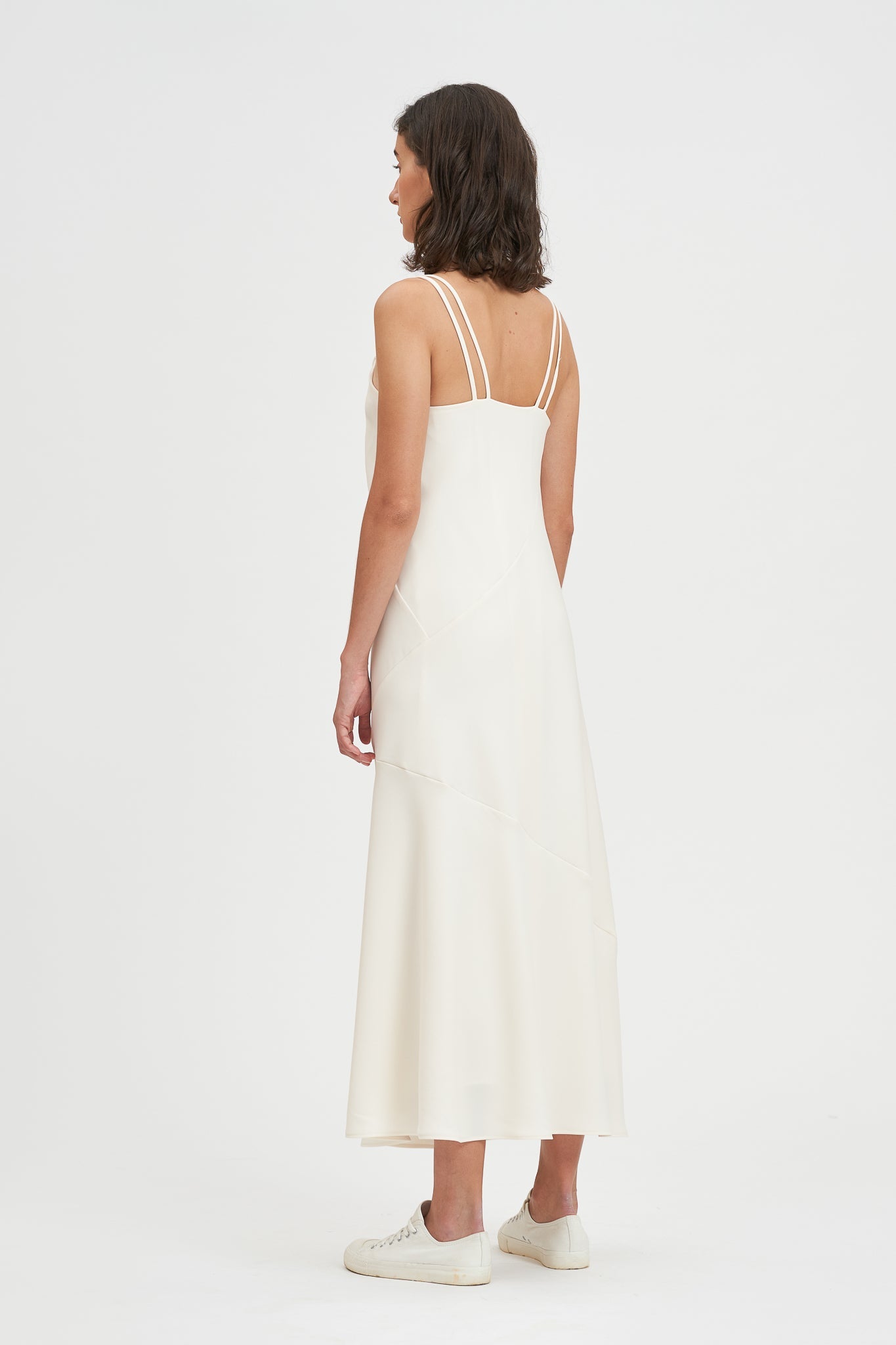 A woman with shoulder length hair is wearing an ankle length white scrappy dress. She is wearing white tennis shoes.