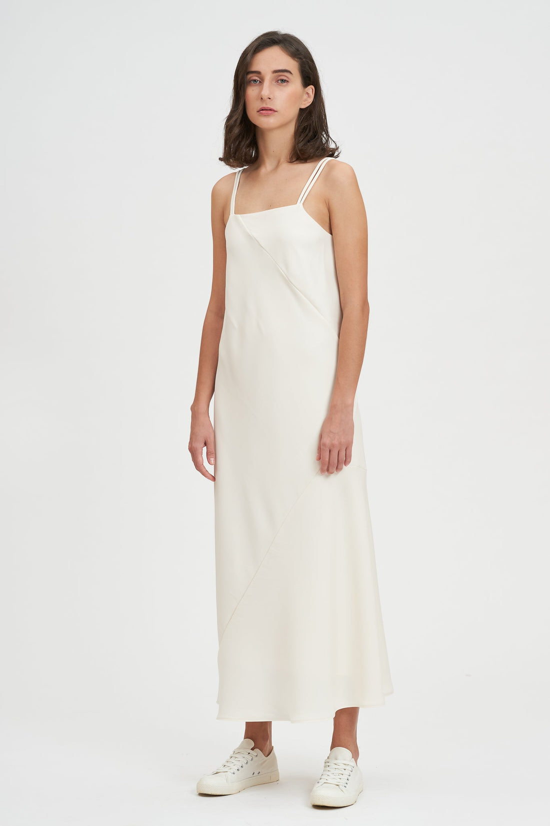 A woman with shoulder length hair is wearing an ankle length white scrappy dress. She is wearing white tennis shoes.