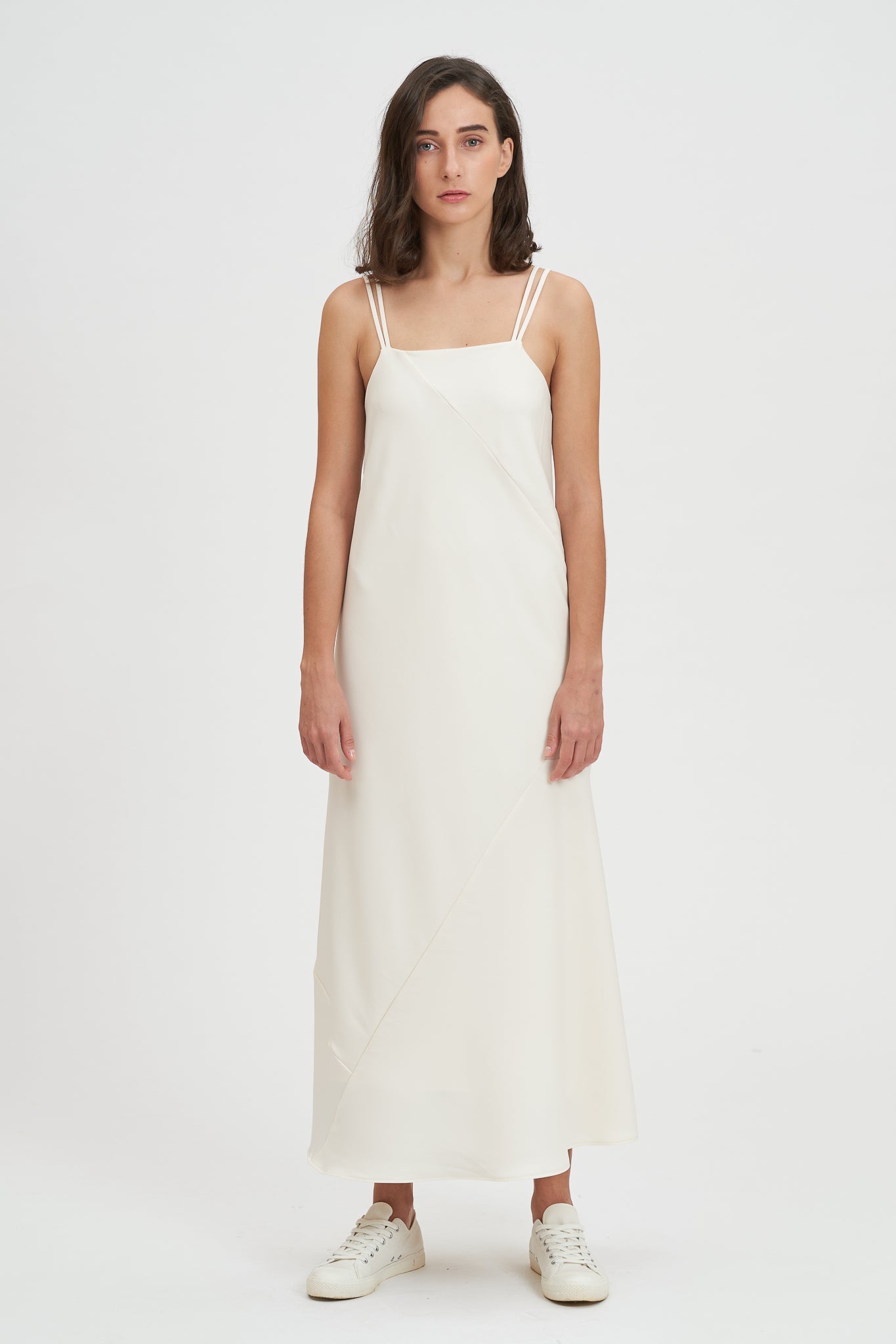 A woman with shoulder length hair is wearing an ankle length white scrappy dress. She is wearing white tennis shoes.