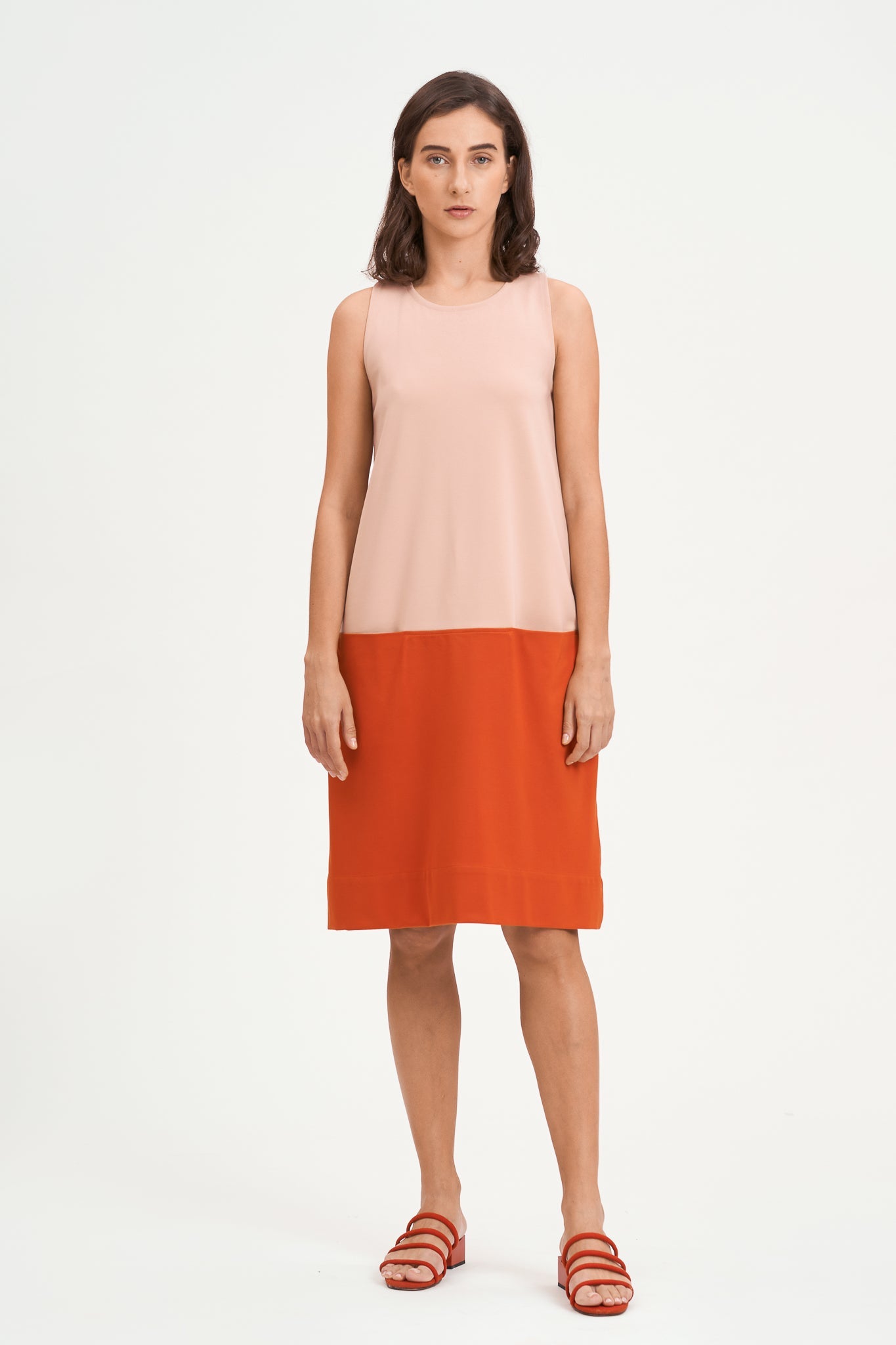 A woman with shoulder length brown hair is wearing a knee length pink and nude dress. She is also wearing red strappy sandals.