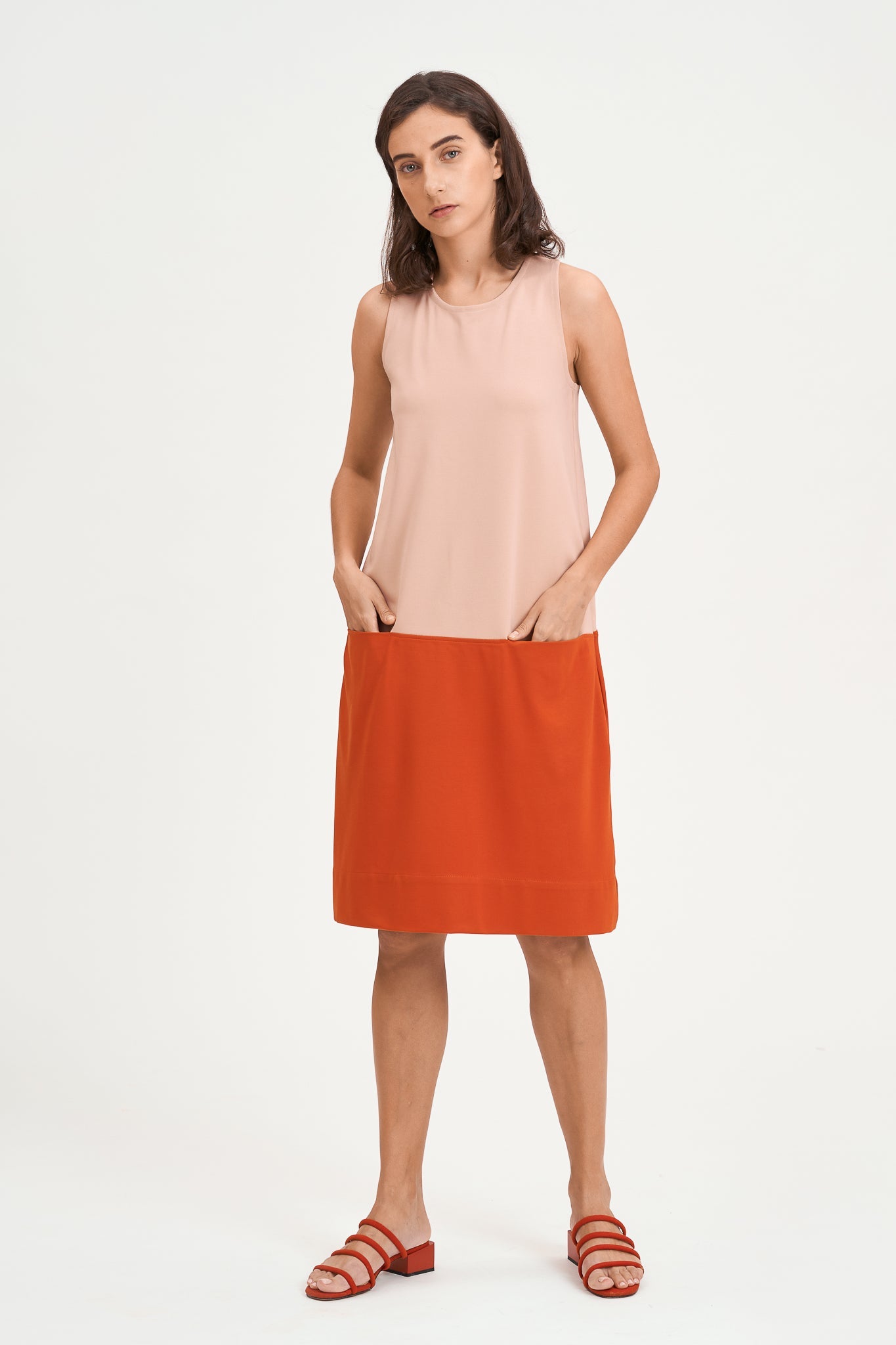 A woman with shoulder length brown hair is wearing a knee length pink and nude dress. She is also wearing red strappy sandals.