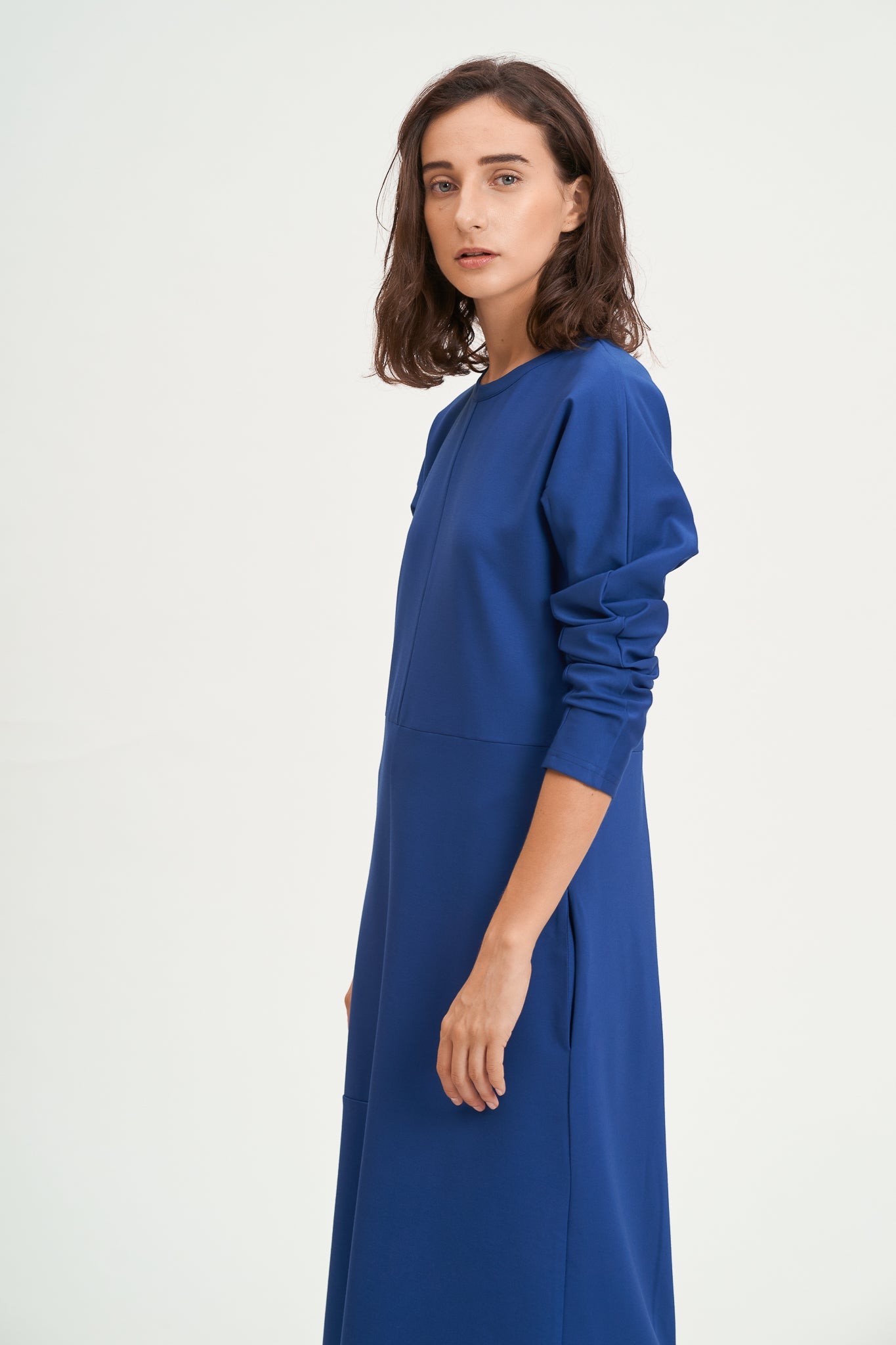 A woman with shoulder length hair is wearing an ankle length blue dress with long sleeves.