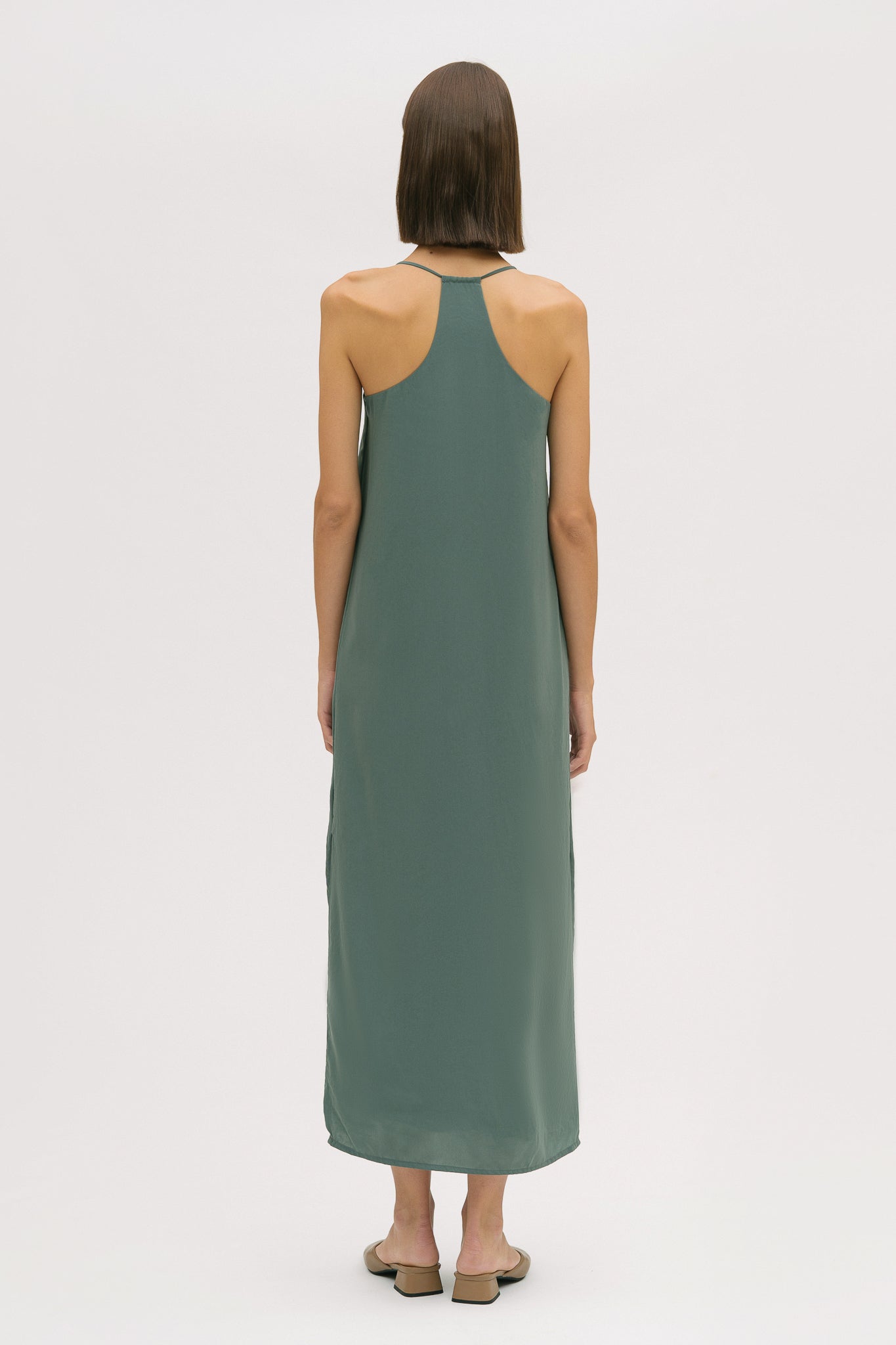 A woman with short brown hair is wearing a long green silky dress with straps. She has beige mules on.