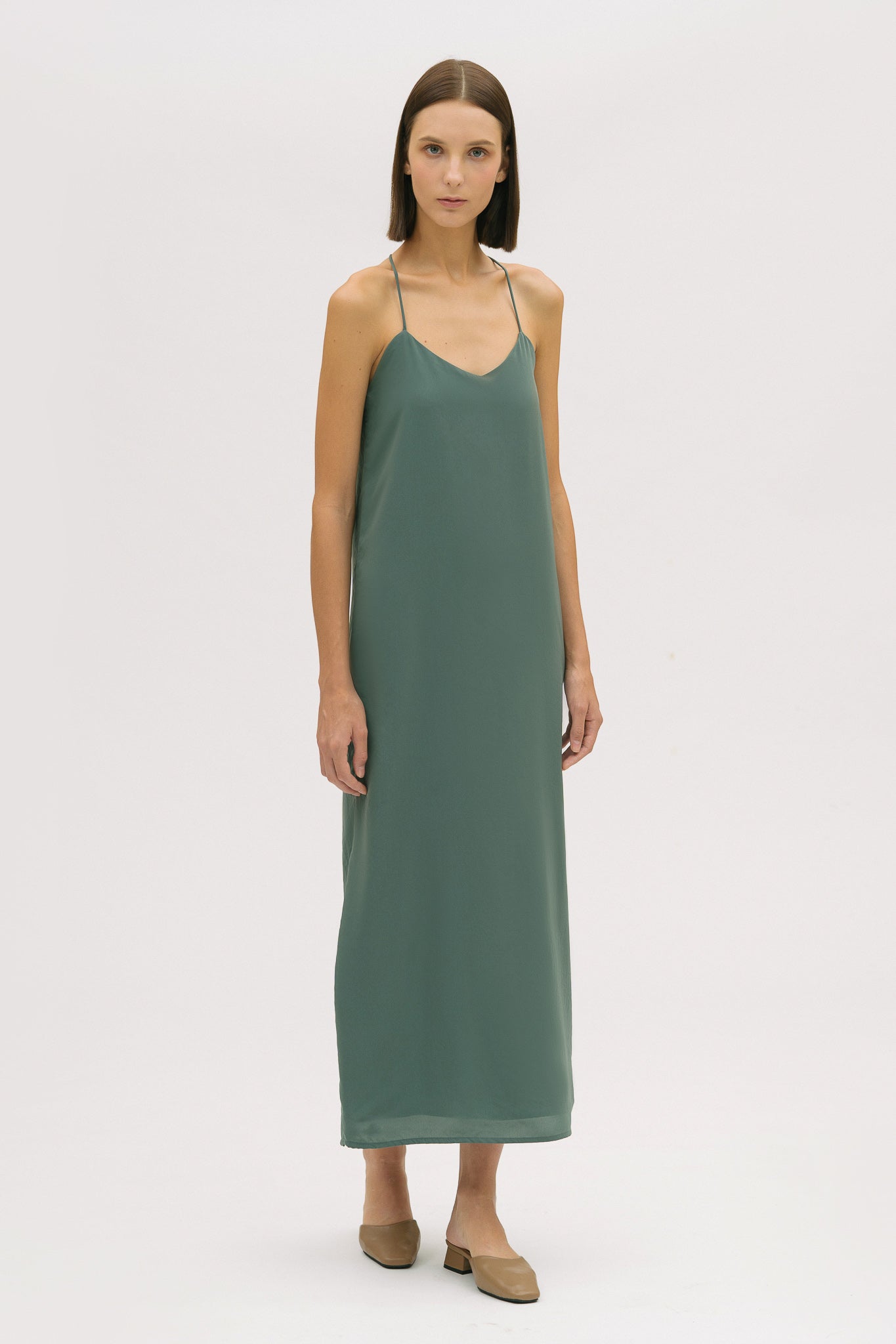 A woman with short brown hair is wearing a long green silky dress with straps. She has beige mules on.
