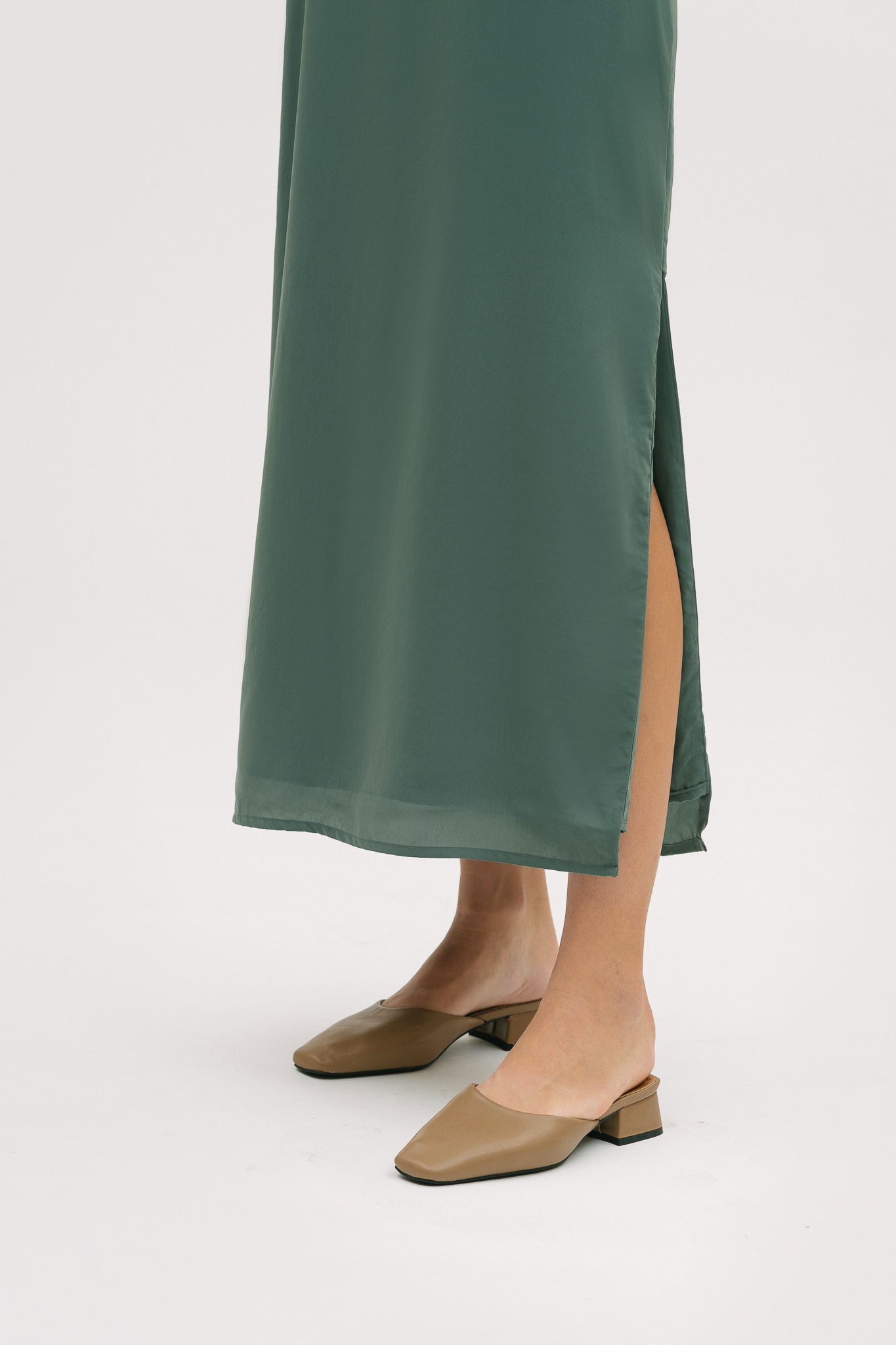 A woman is wearing a long green silky dress with straps. She has beige mules on.