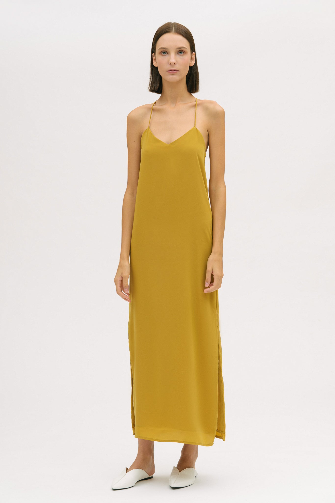 A woman with short brown hair is wearing a long yellow silky dress with straps. She has white mules on.