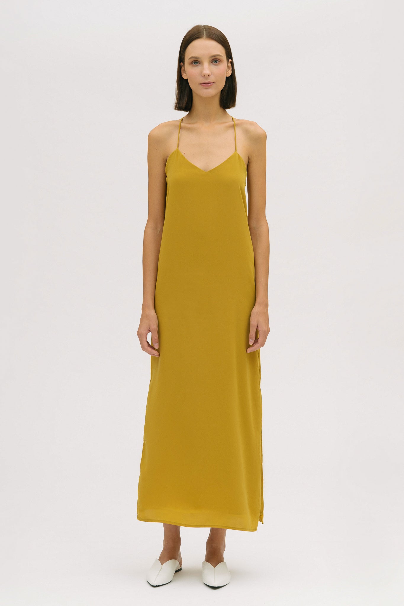A woman with short brown hair is wearing a long yellow silky dress with straps. She has white mules on.