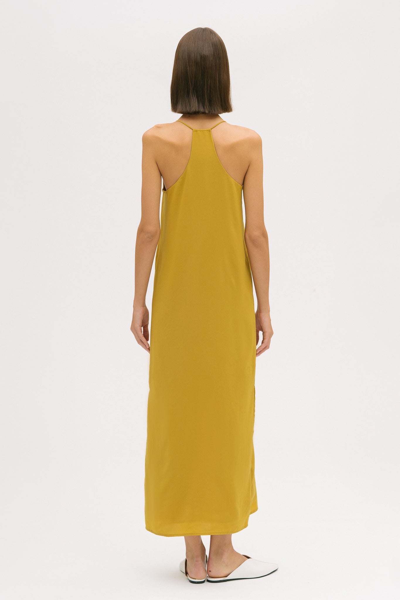 A woman with short brown hair is wearing a long yellow silky dress with straps. She has white mules on.