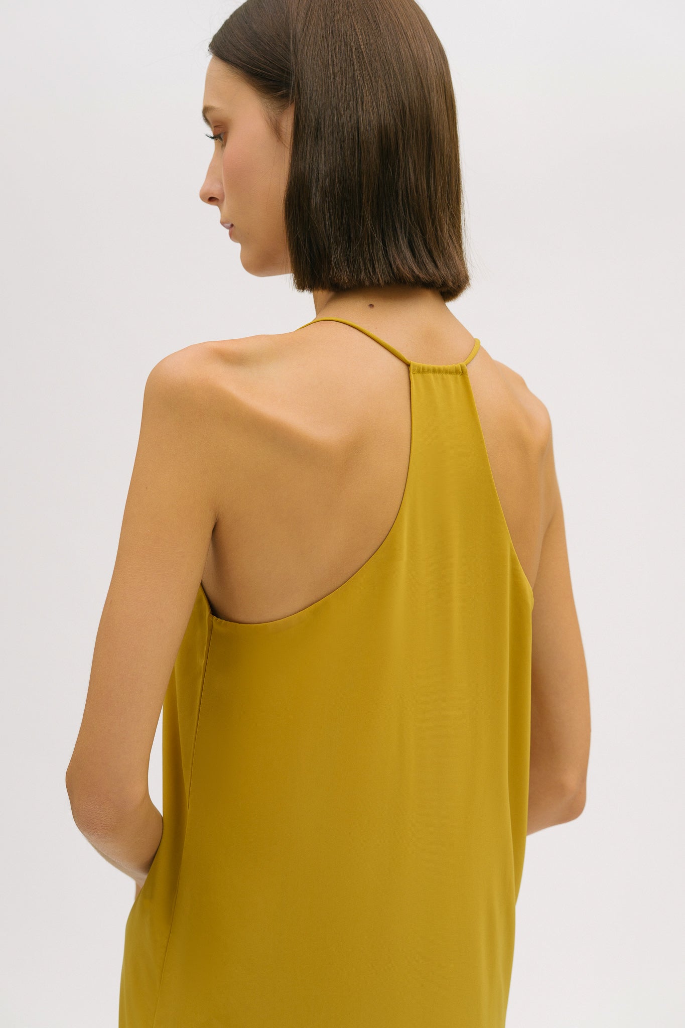 A woman with short brown hair is wearing a long yellow silky dress with straps. She has white mules on.
