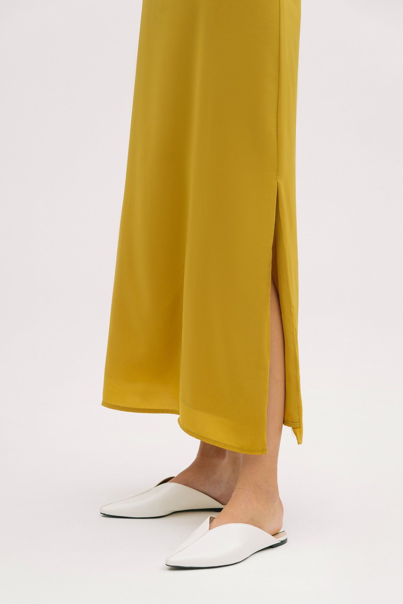 A woman with short brown hair is wearing a long yellow silky dress with straps. She has white mules on.