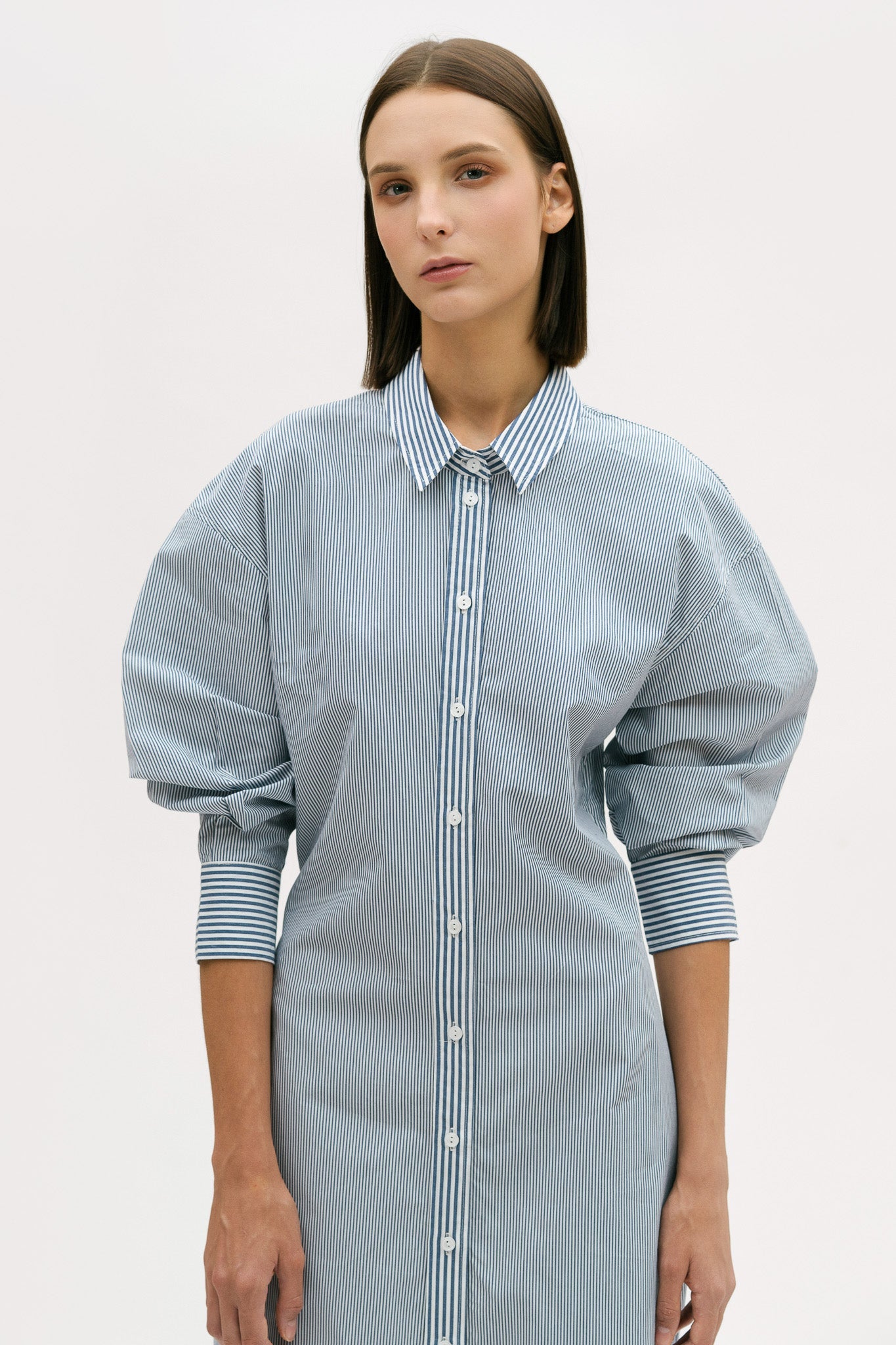 A woman with short brown hair is wearing a knee length blue and white pin striped shirt dress.