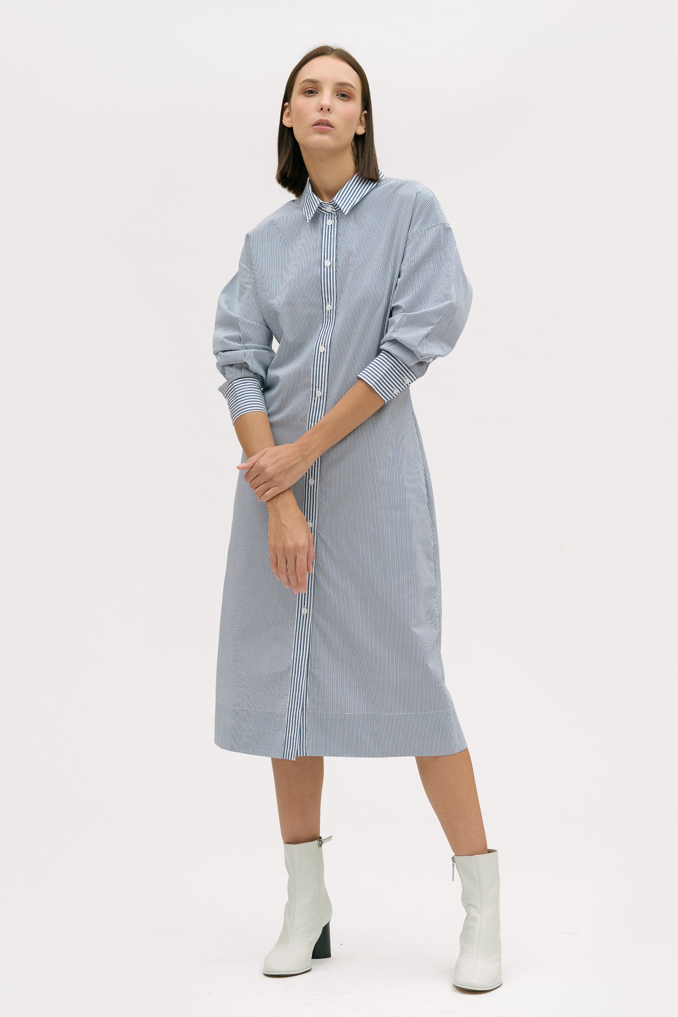 A woman with short brown hair is wearing a knee length blue and white pin striped shirt dress. She is wearing white ankle boots.