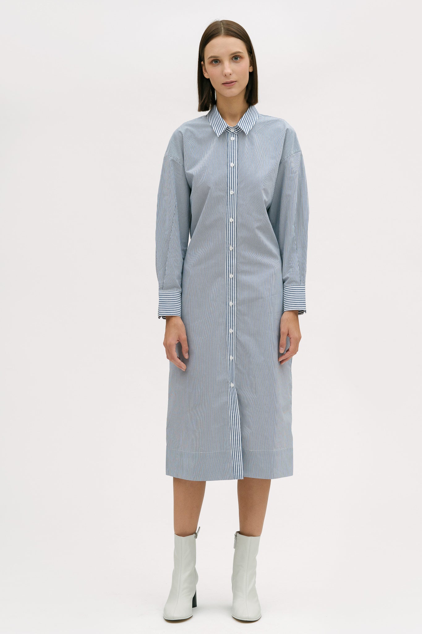 A woman with short brown hair is wearing a knee length blue and white pin striped shirt dress. She is wearing white ankle boots.