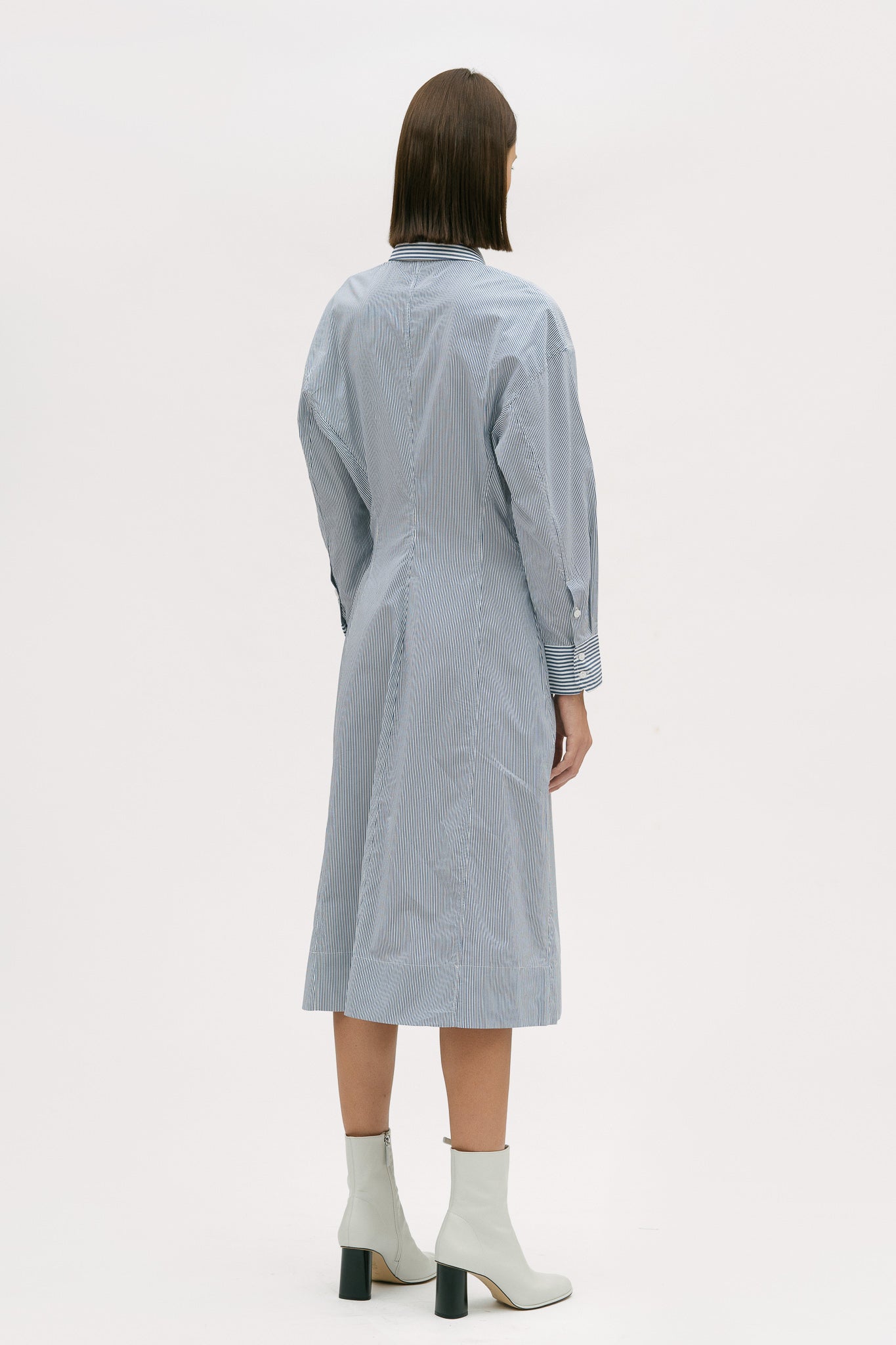 A woman with short brown hair is wearing a knee length blue and white pin striped shirt dress. She is wearing white ankle boots.
