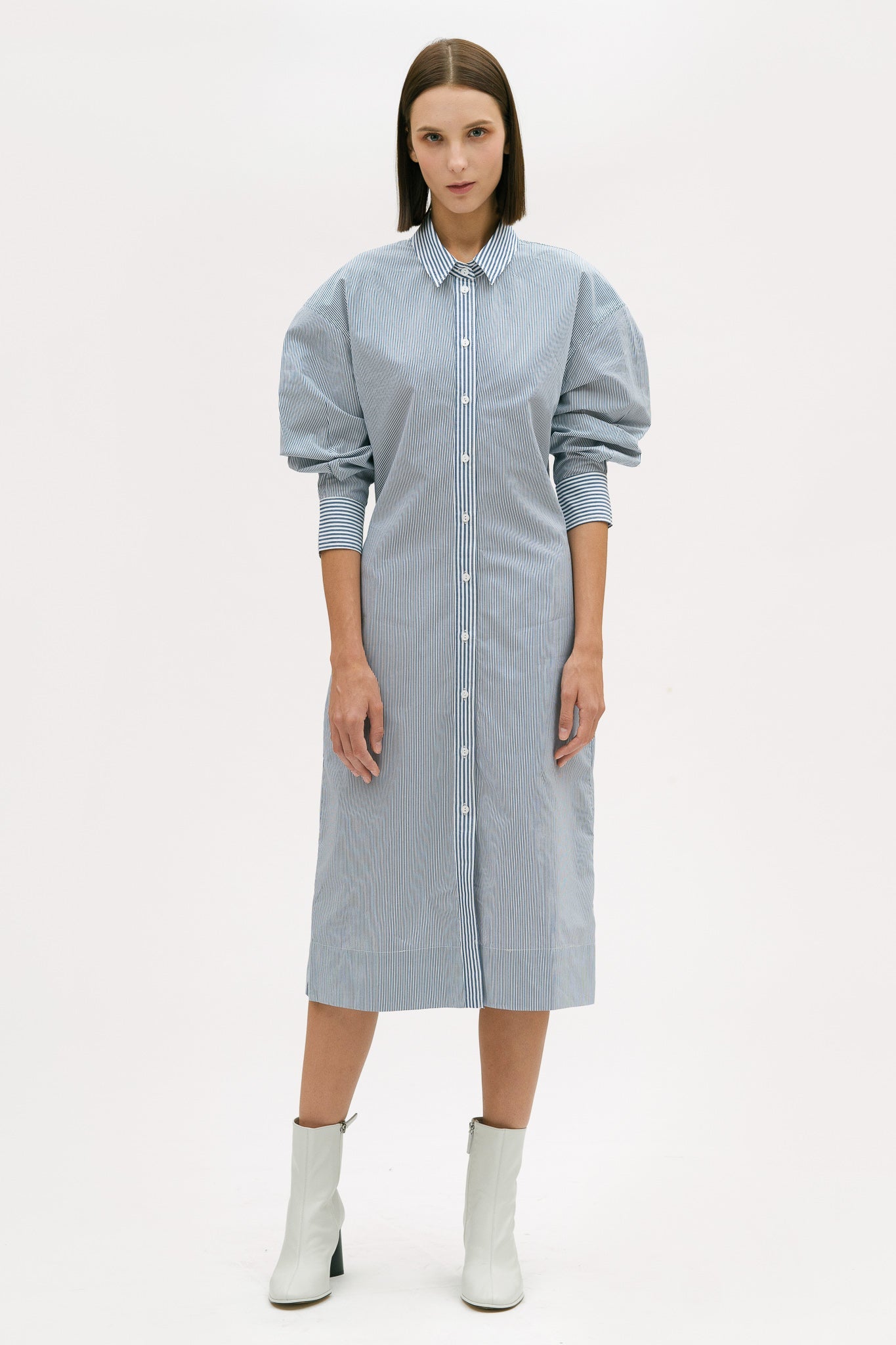 A woman with short brown hair is wearing a knee length blue and white pin striped shirt dress. She is wearing white ankle boots.