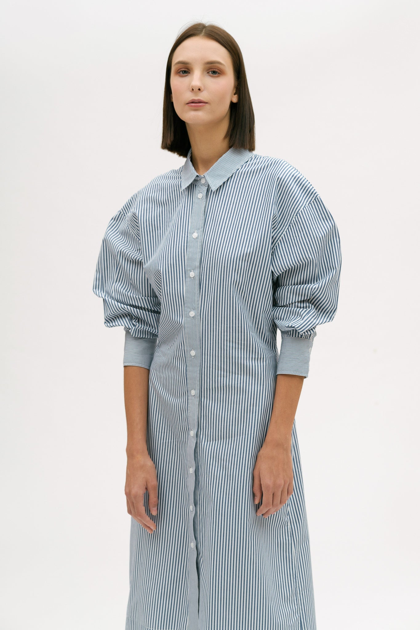 A woman with short brown hair is wearing a knee length blue and white pin striped shirt dress.