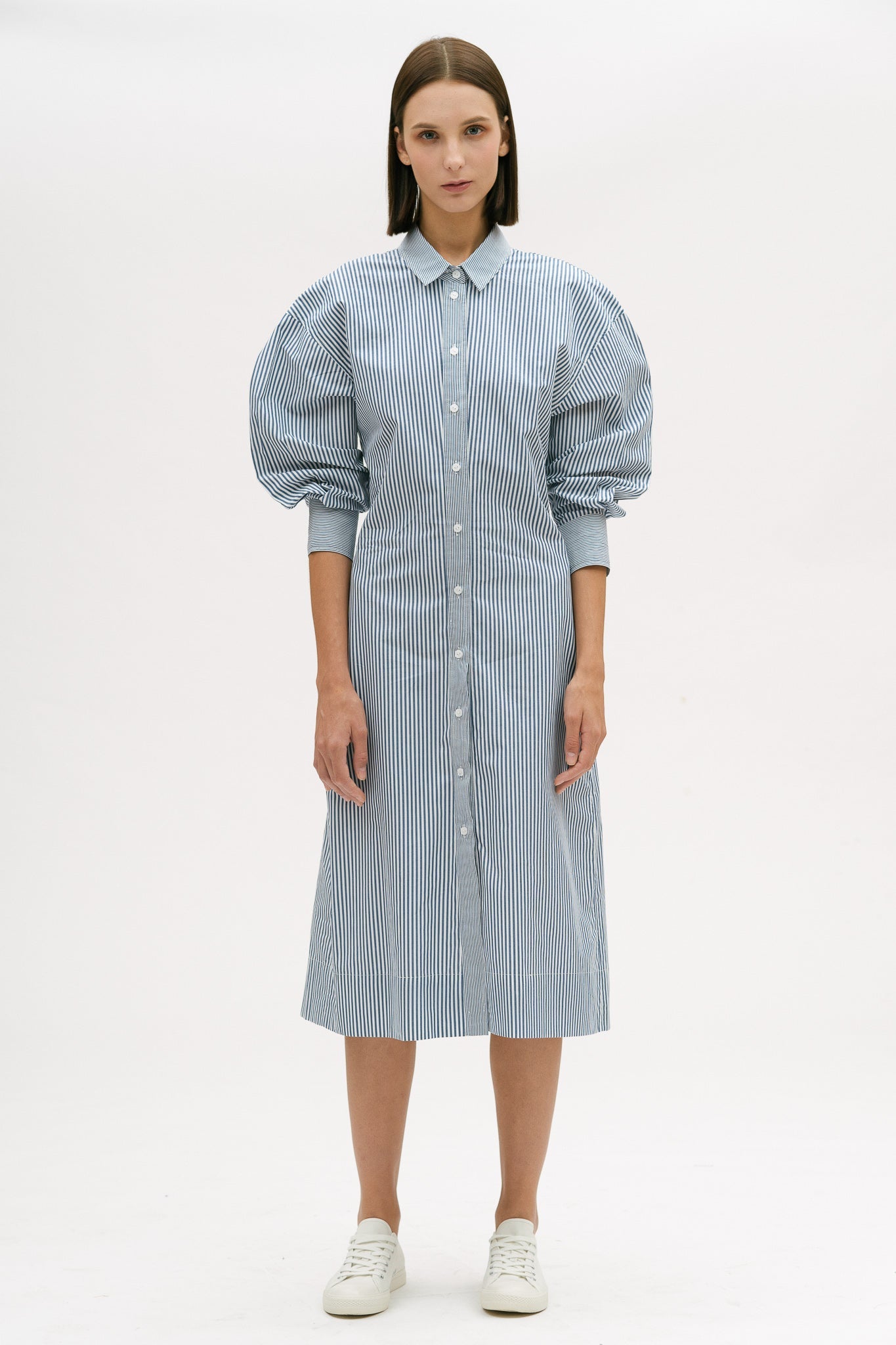 A woman with short brown hair is wearing a knee length blue and white pin striped shirt dress. She is wearing white tennis shoes.