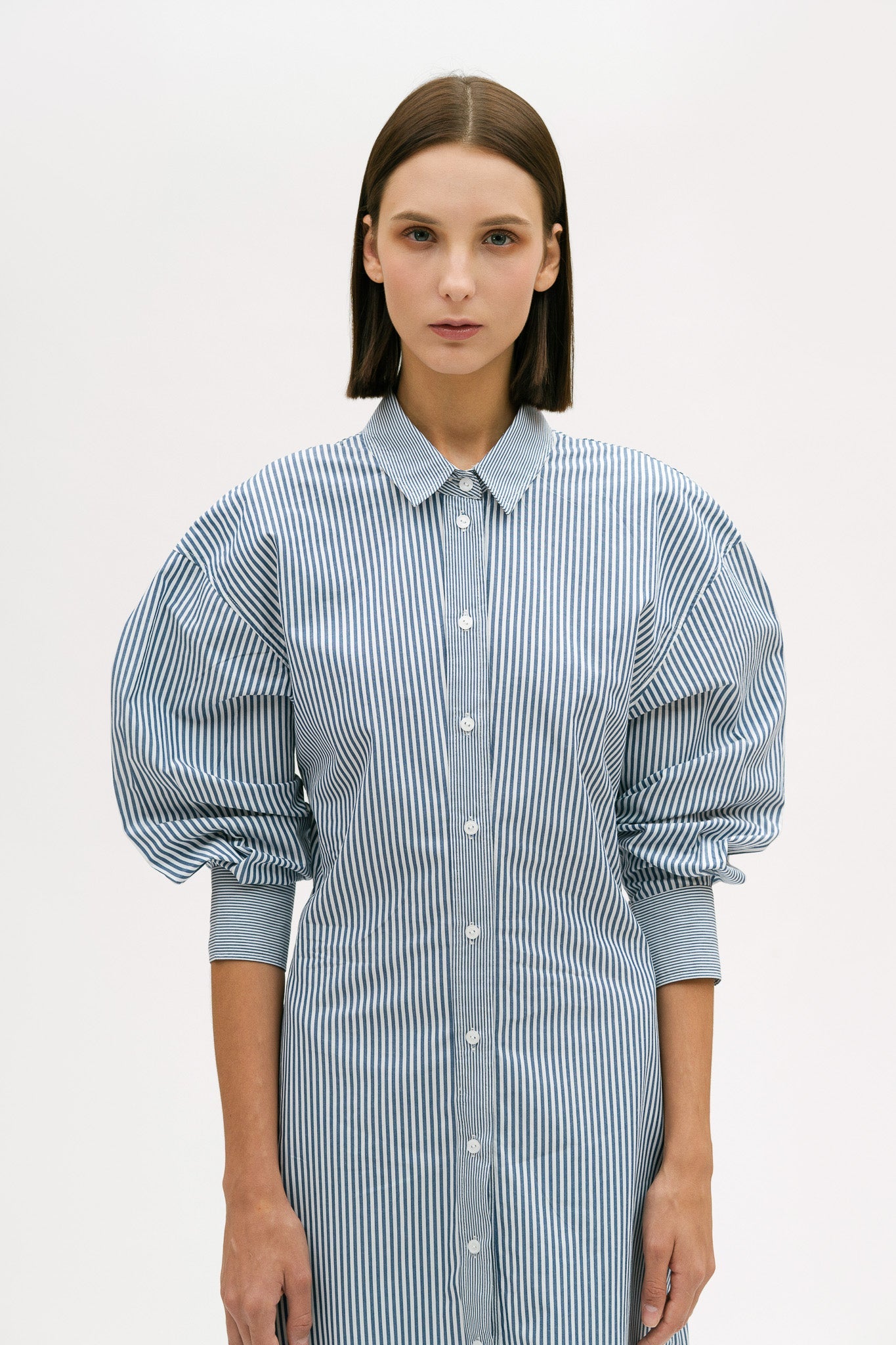 A woman with short brown hair is wearing a knee length blue and white pin striped shirt dress.