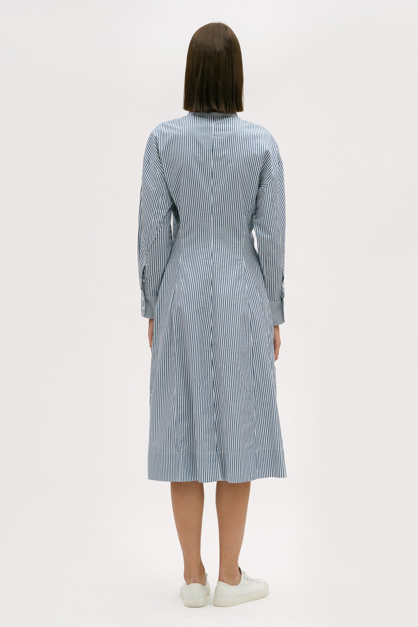 A woman with short brown hair is wearing a knee length blue and white pin striped shirt dress. She is wearing white tennis shoes.