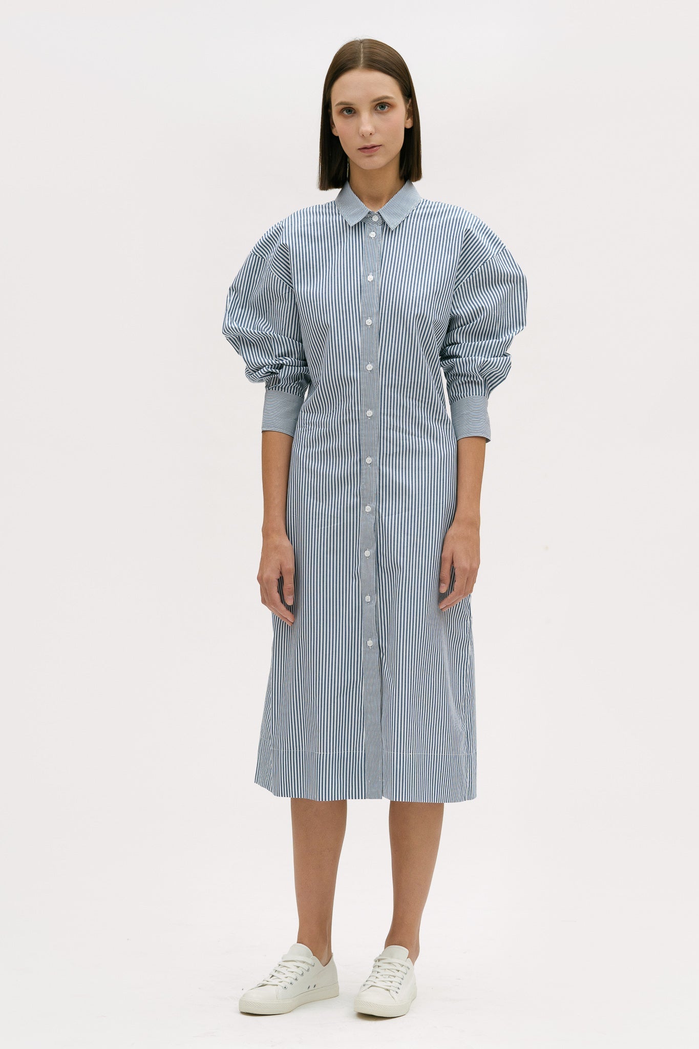 A woman with short brown hair is wearing a knee length blue and white pin striped shirt dress. She is wearing white tennis shoes.