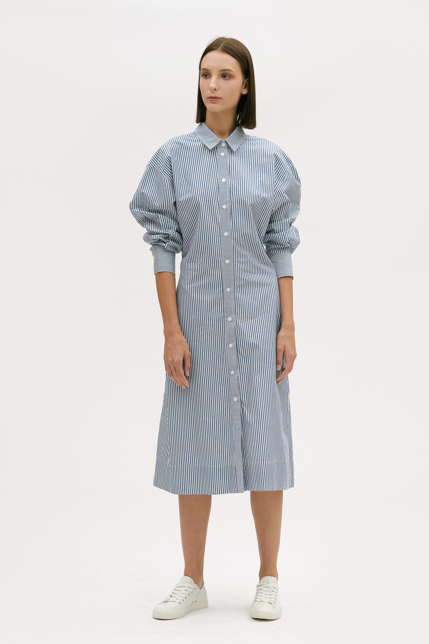A woman with short brown hair is wearing a knee length blue and white pin striped shirt dress. She is wearing white tennis shoes.