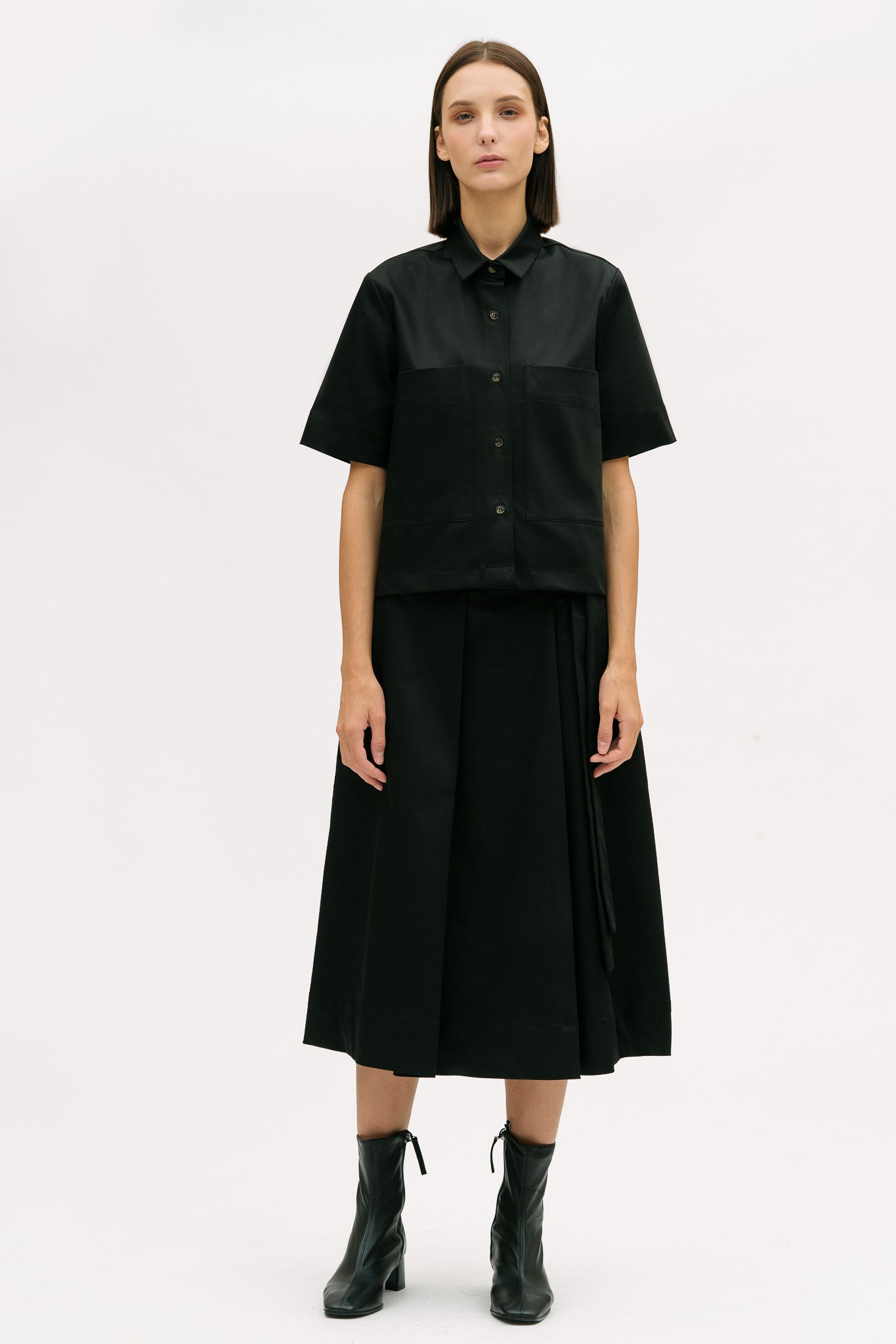 A woman is wearing a black shirt and black calf length skirt. She is also wearing black boots
