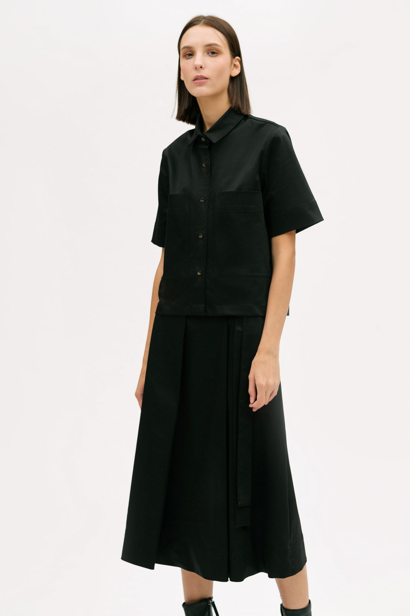 A woman with shoulder length brown hair is wearing a black utility shirt, a long black skirt and black boots
