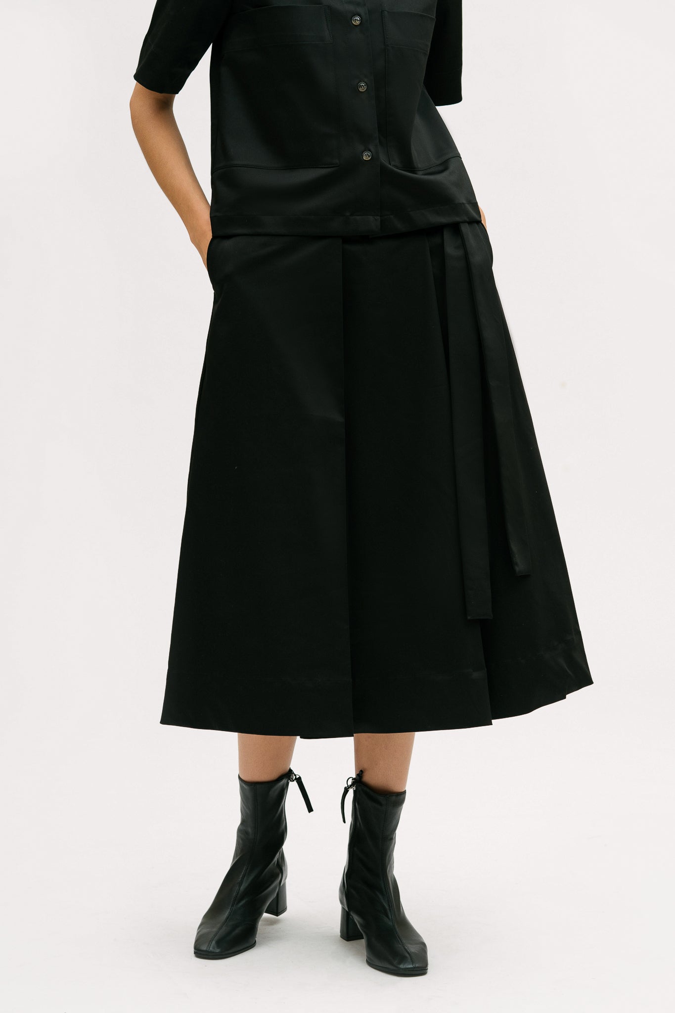 A woman is wearing a black shirt and black calf length skirt. She is also wearing black boots