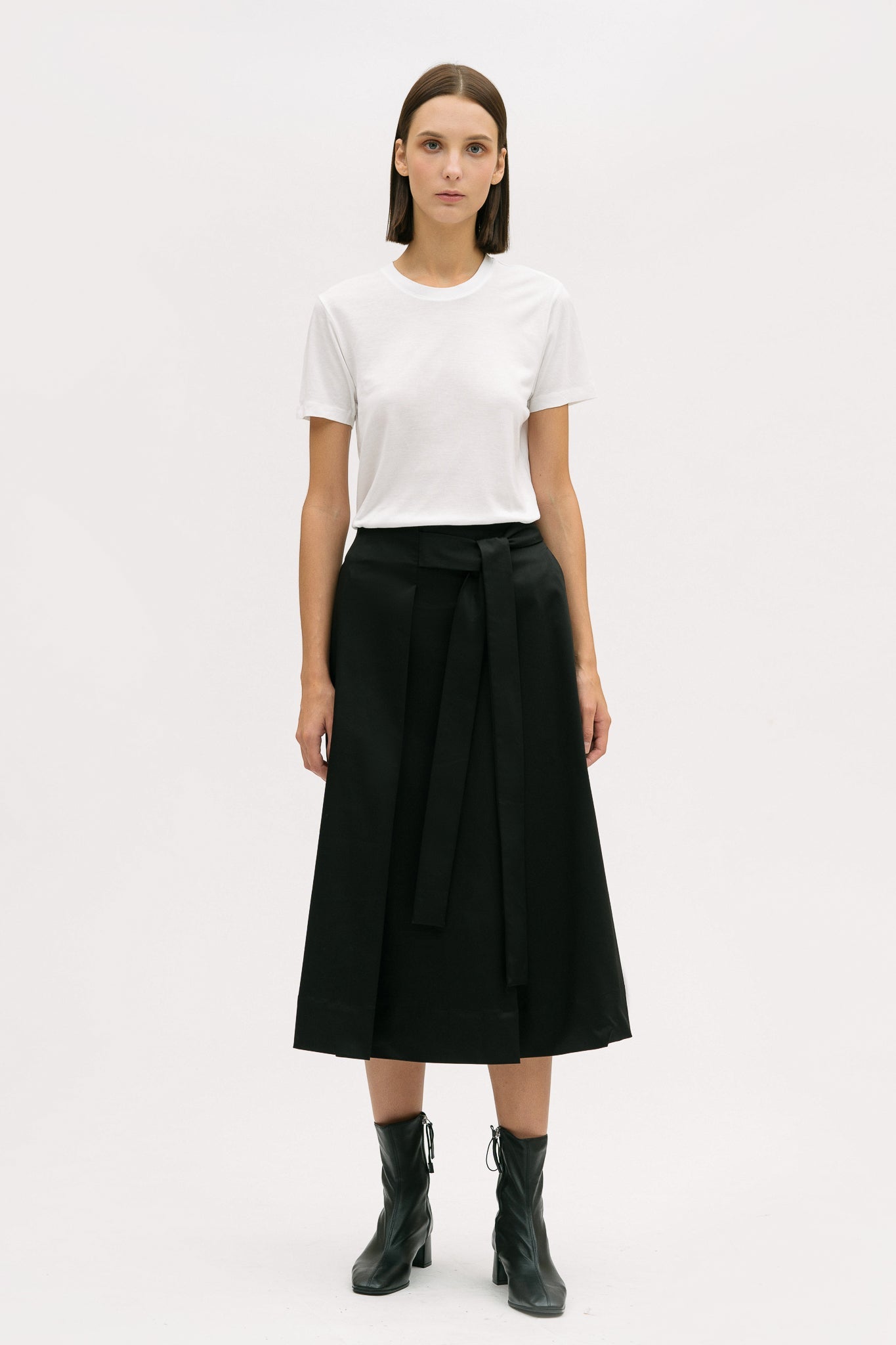 A woman is wearing a white t shirt and black calf length skirt. She is also wearing black boots