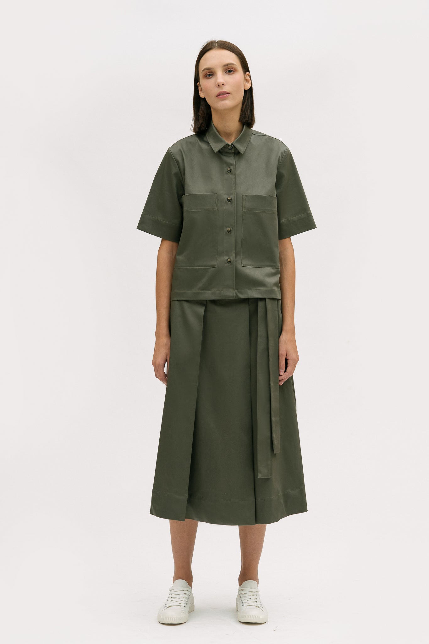 A woman with shoulder length brown hair is wearing a green shirt and calf length skirt. She is also wearing white tennis shoes.