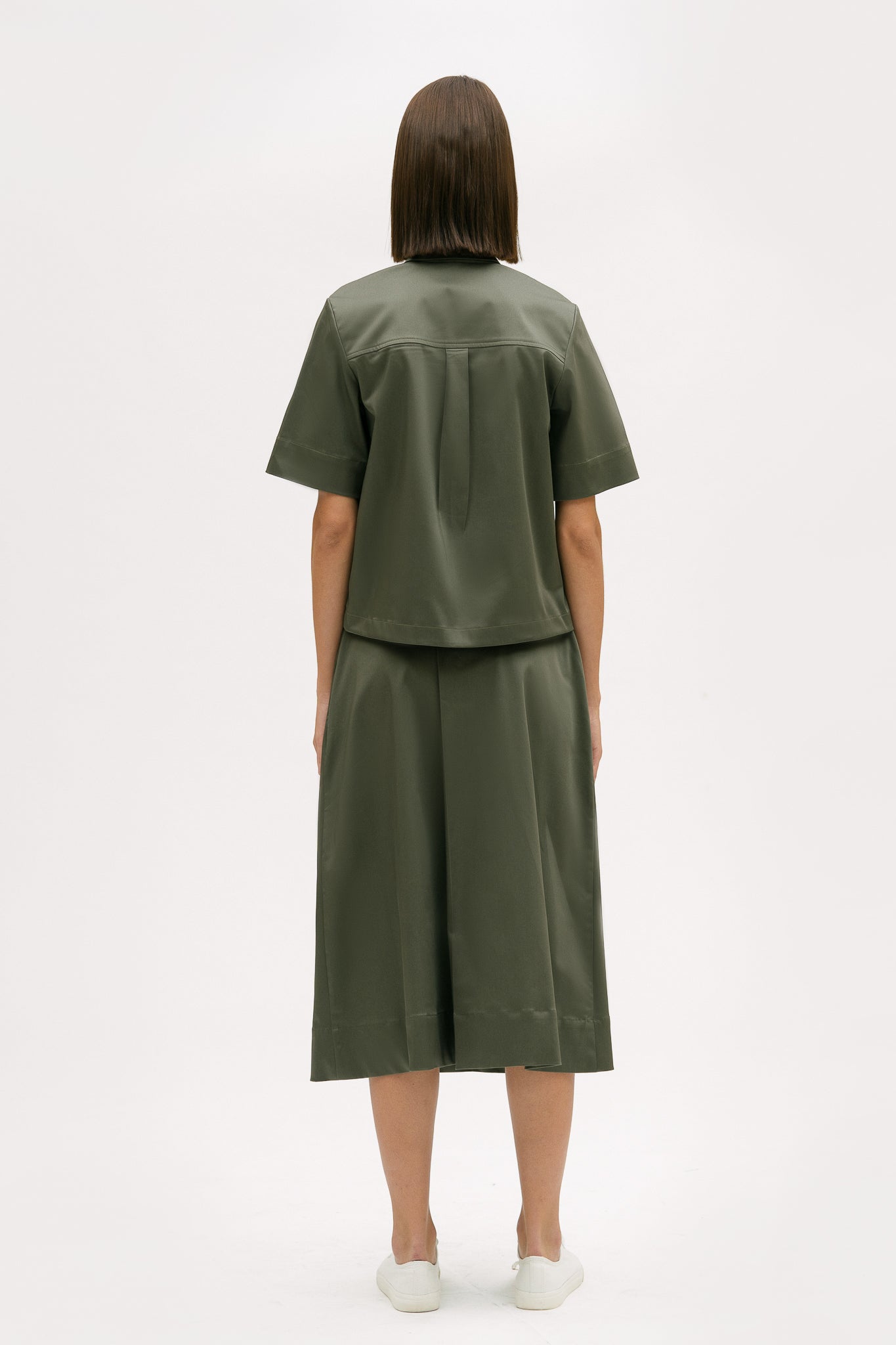 A woman with shoulder length brown hair is wearing a green shirt and calf length skirt. She is also wearing white tennis shoes.