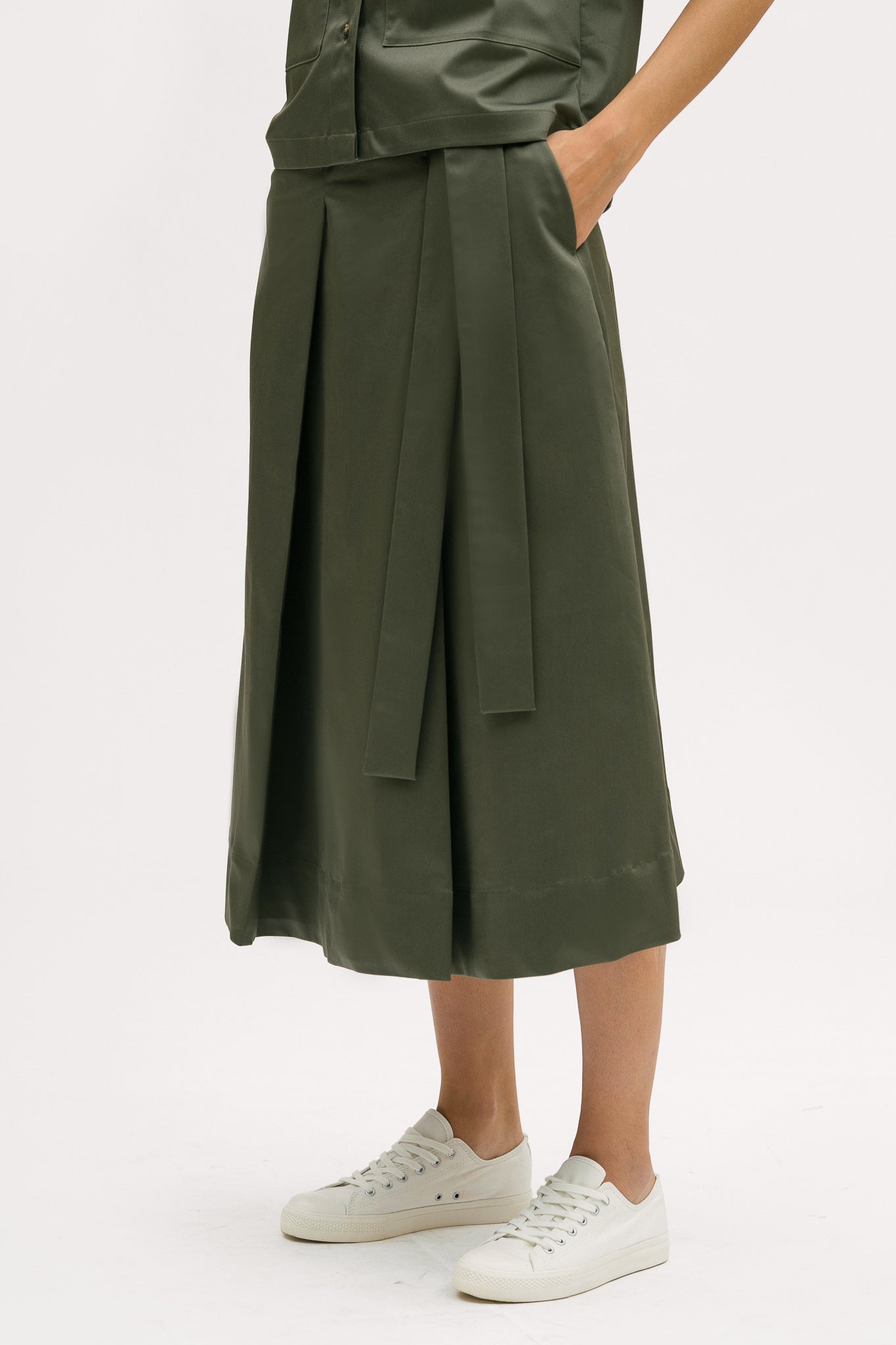 A woman with shoulder length brown hair is wearing a green shirt and calf length skirt. She is also wearing white tennis shoes.