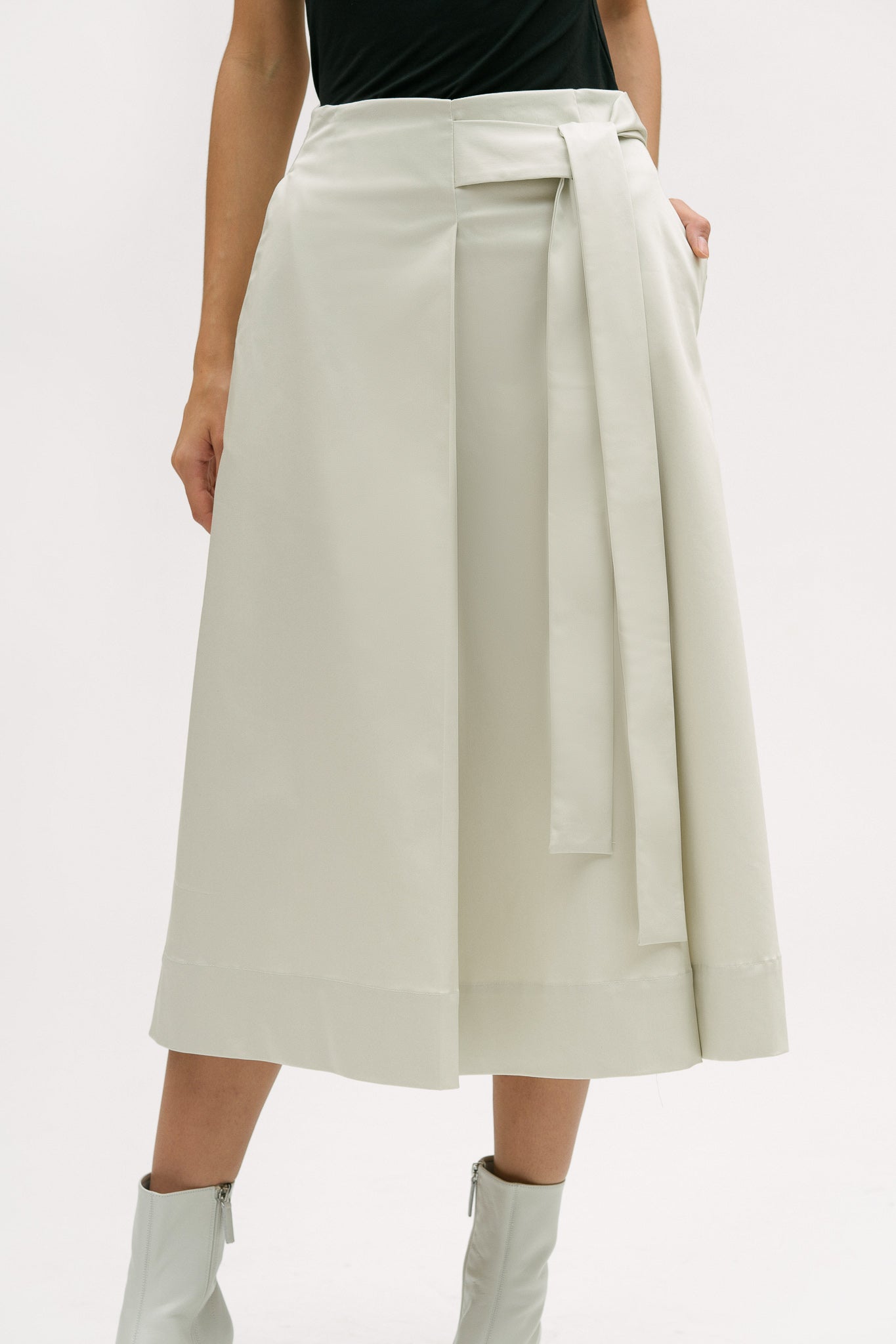 A woman with shoulder length brown hair is wearing a black top and off-white calf length skirt. She is also wearing white boots.