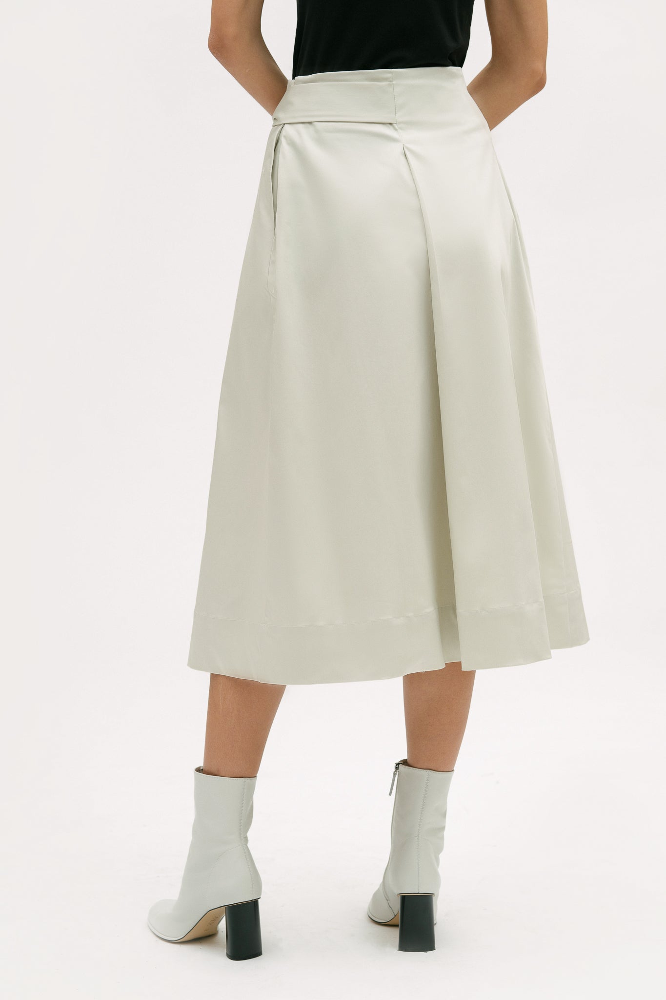 A woman with shoulder length brown hair is wearing a black top and off-white calf length skirt. She is also wearing white boots.