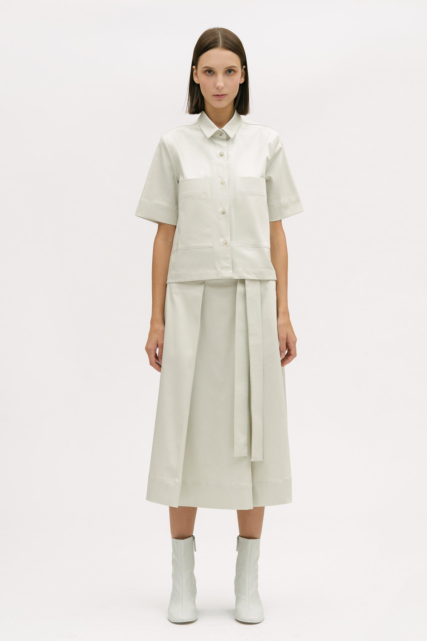 A woman with shoulder length brown hair is wearing an off-white shirt and calf length skirt. She is also wearing white boots.