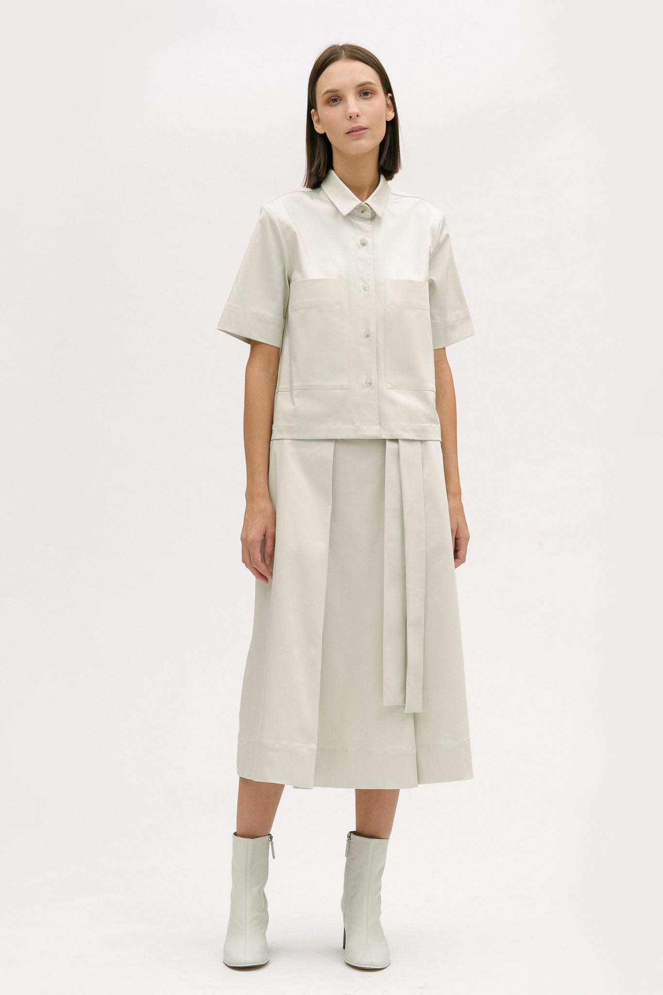 A woman with shoulder length brown hair is wearing an off-white shirt and calf length skirt. She is also wearing white boots.