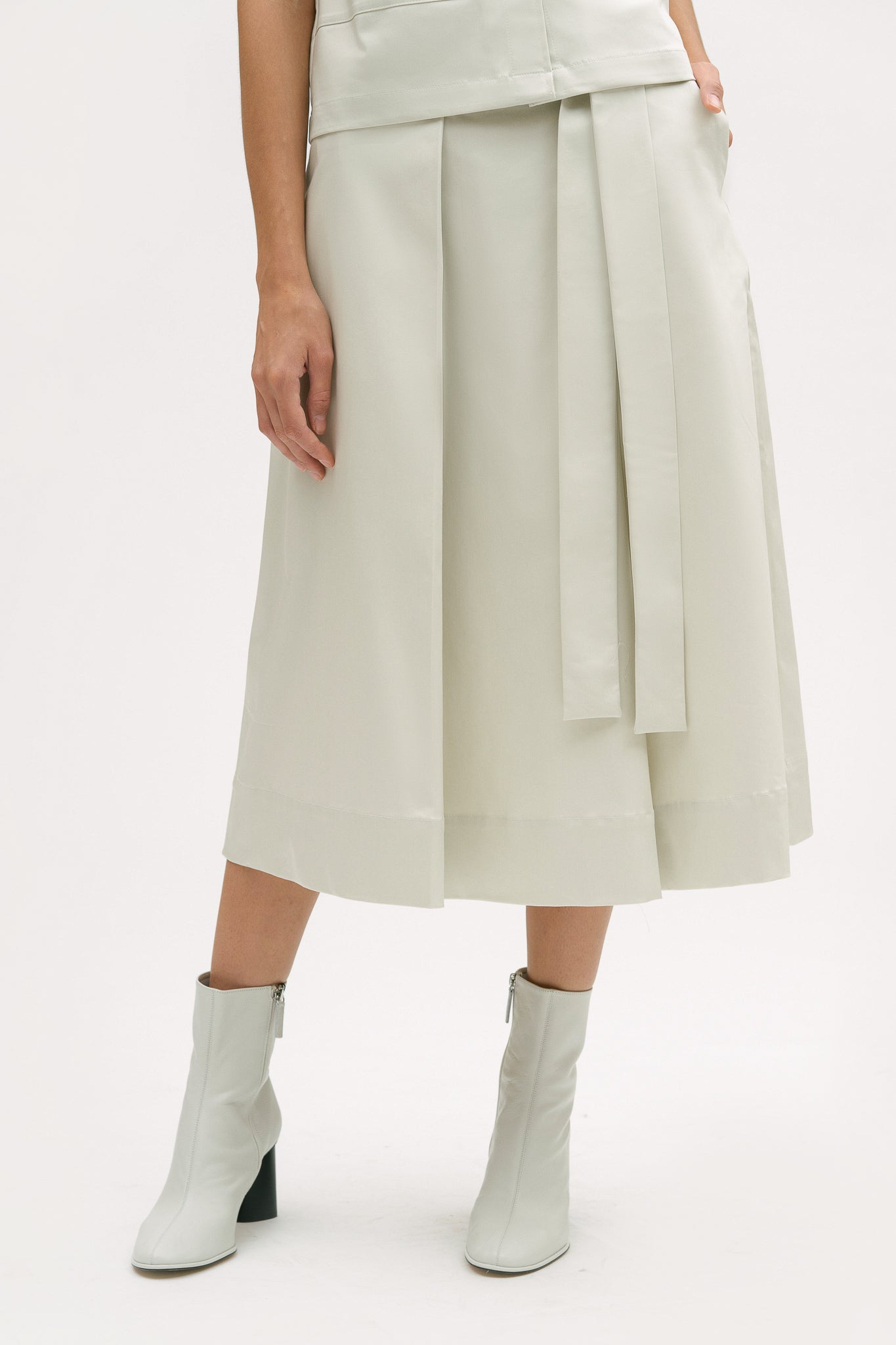 A woman with shoulder length brown hair is wearing an off-white shirt and calf length skirt. She is also wearing white boots.